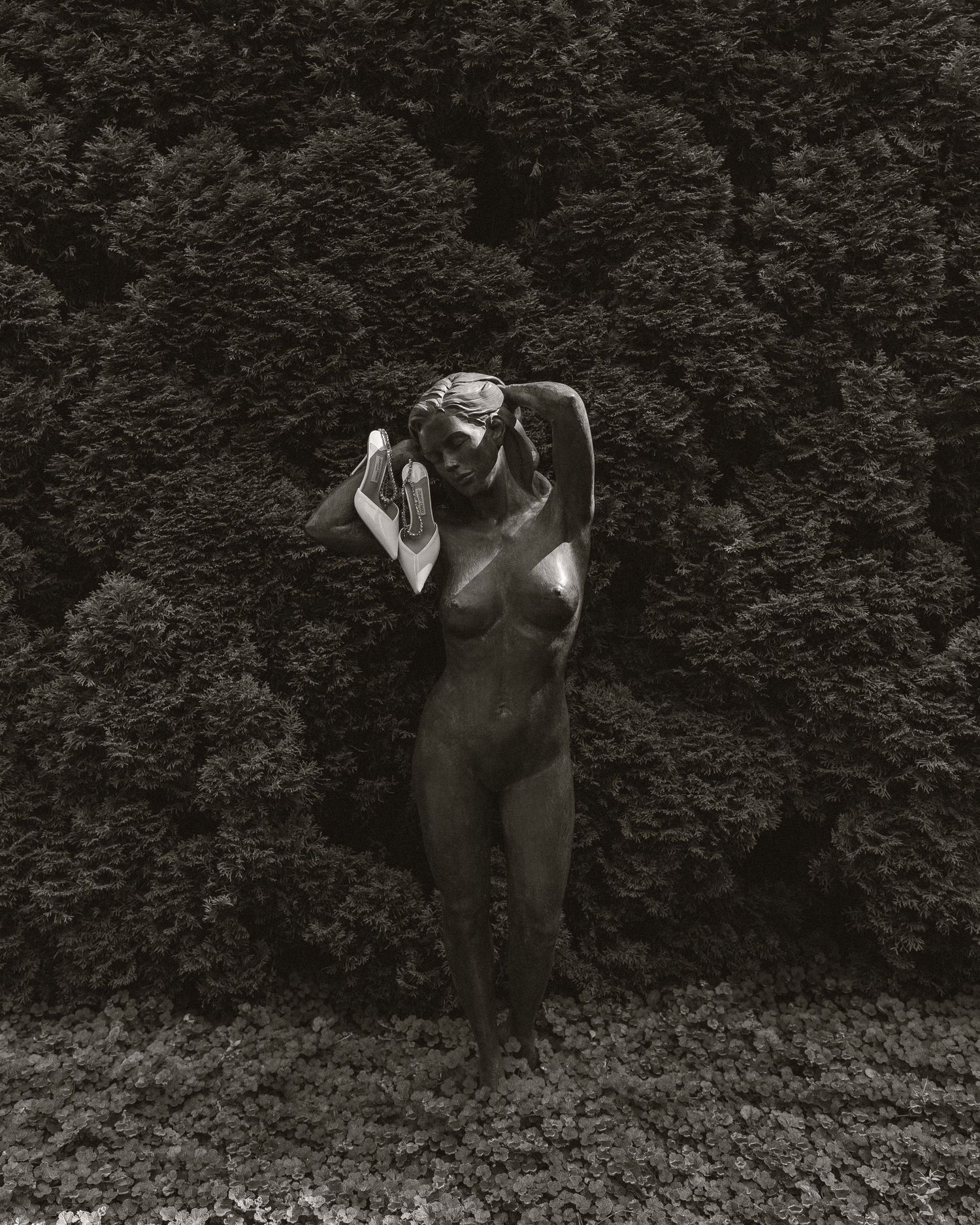 Black and white photograph of a bronze female nude sculpture standing against a lush dense hedge in a garden.