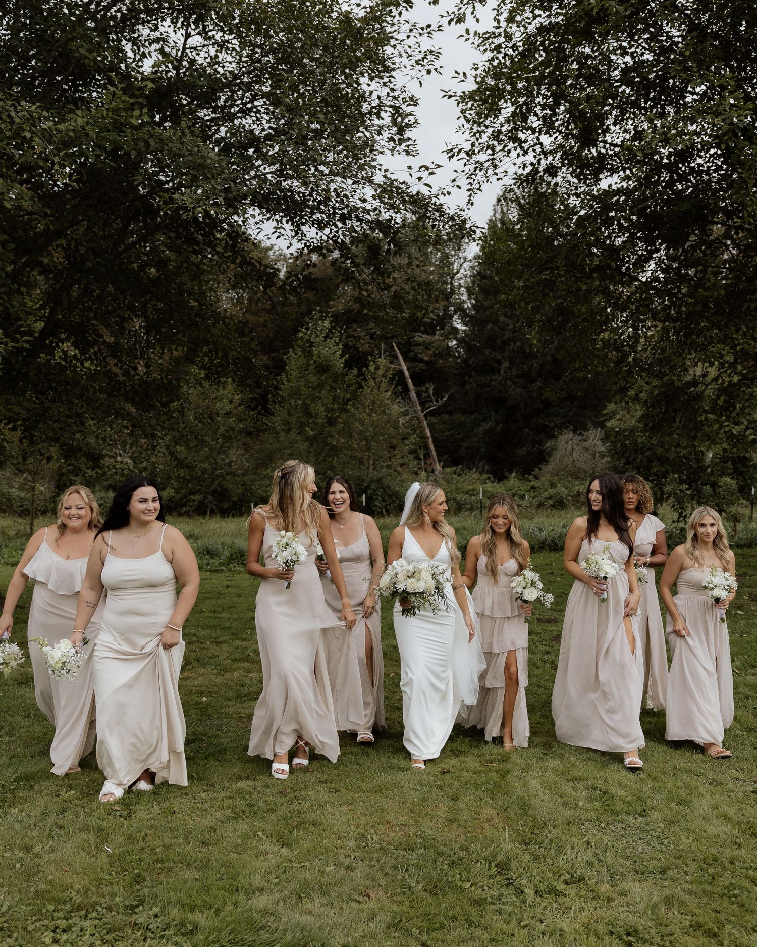 Wedding party walking and laughing together on grassy field with trees in background.