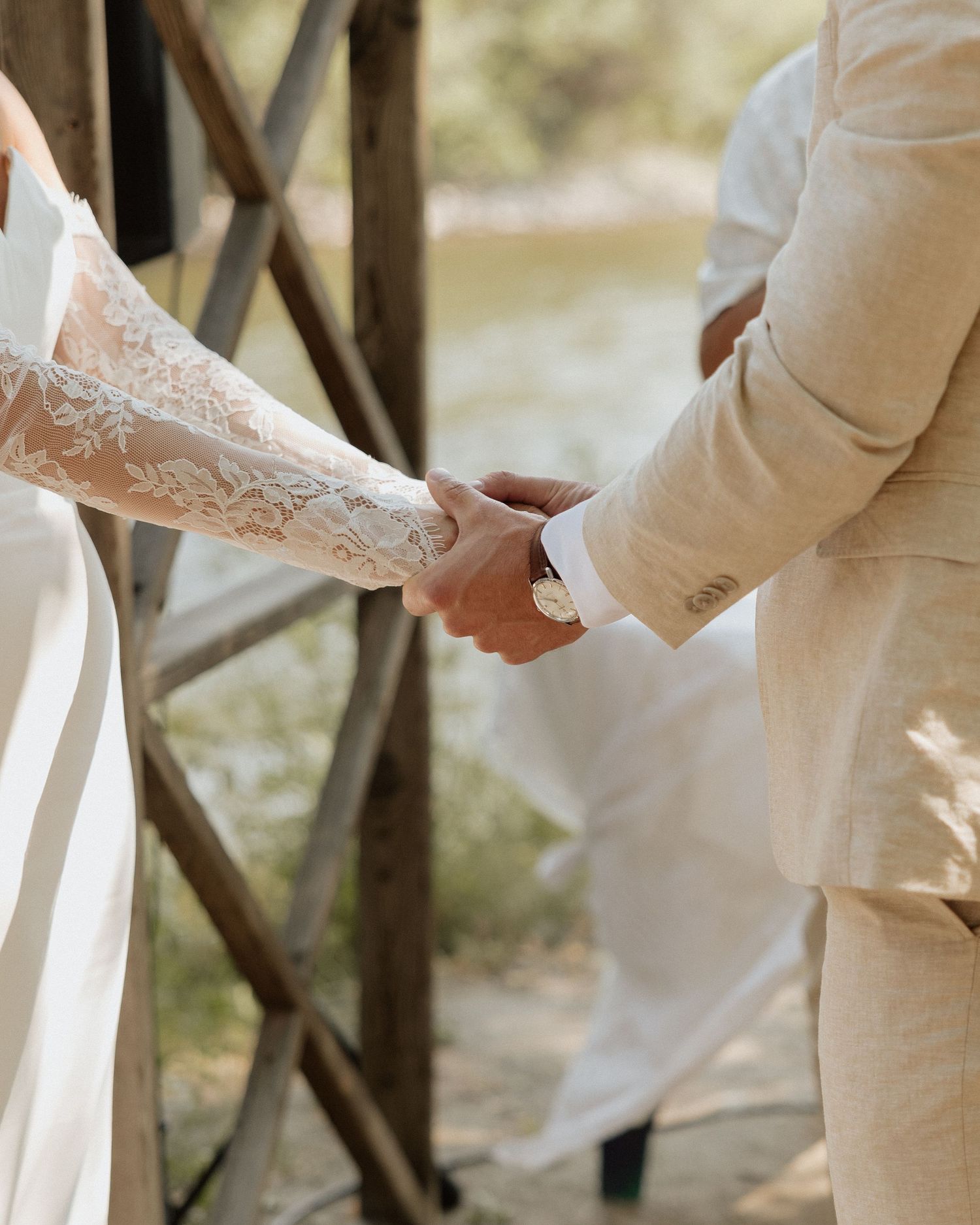 A close-up of hands clasped together during a wedding ceremony, showing delicate lace sleeves and elegant formal attire.