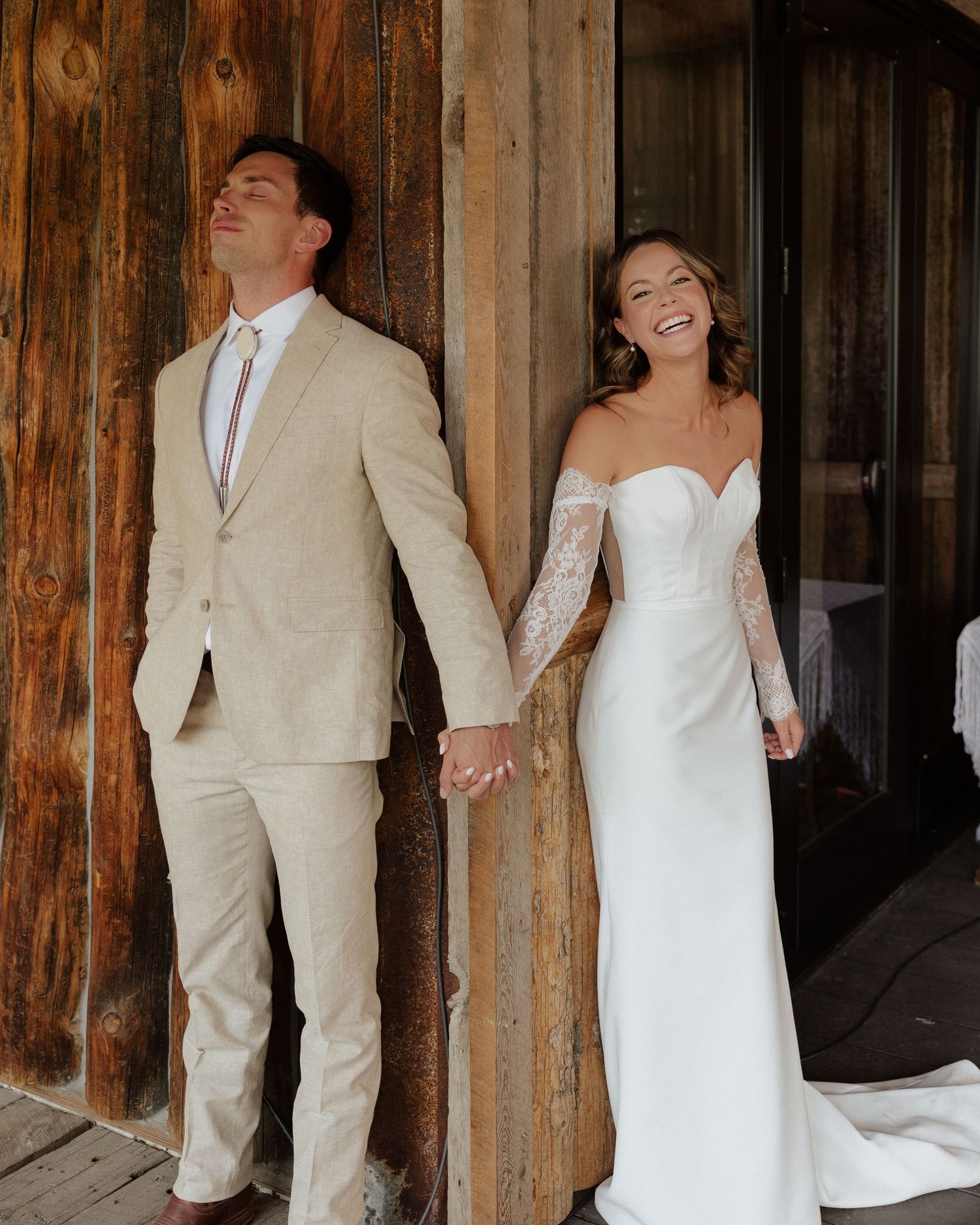 Joyful couple shares laughter during first look photos against wooden wall in wedding attire.