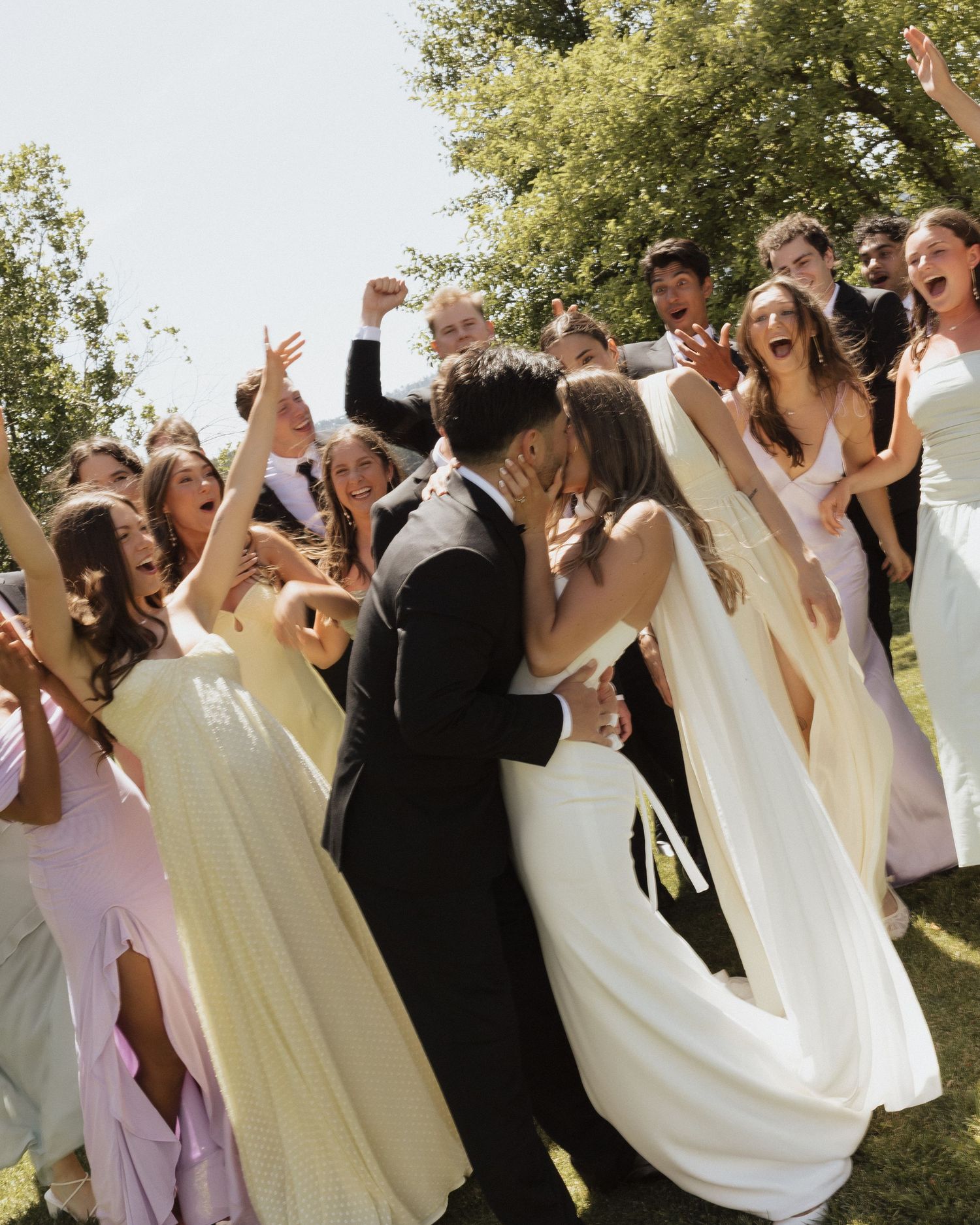 A wedding party dances and celebrates joyfully outdoors on a sunny day wearing white and lilac attire.