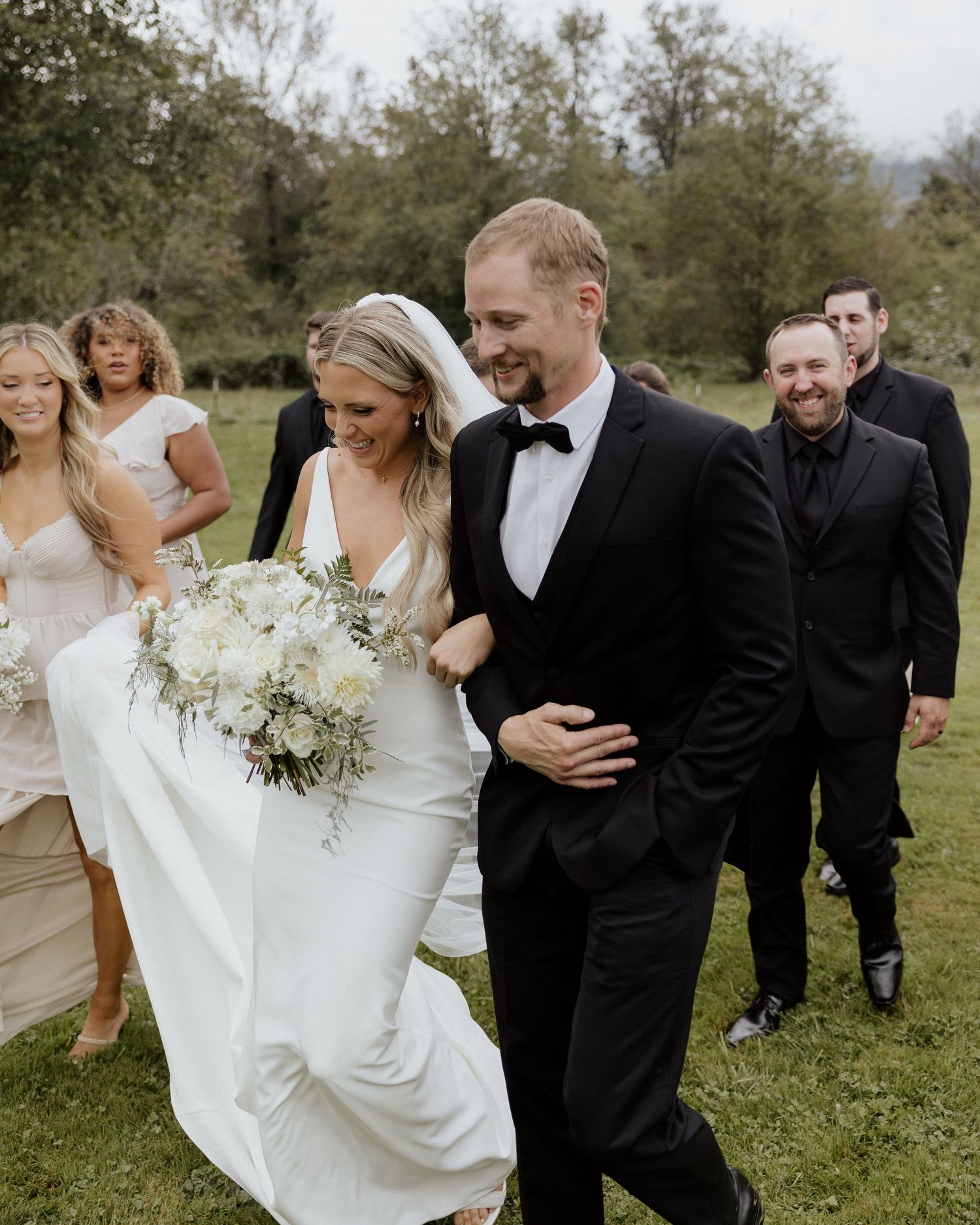 A bride in a flowing white dress walks with her partner across a grassy field as their wedding party follows behind.