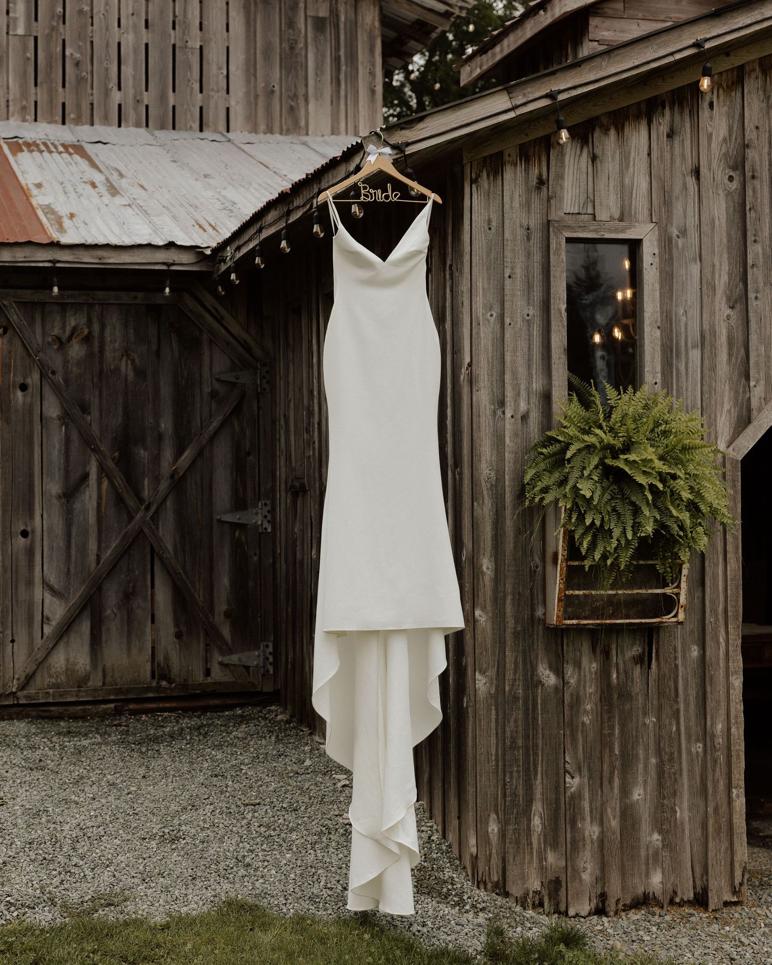 Elegant white wedding dress hanging on rustic barn exterior with fern decoration.