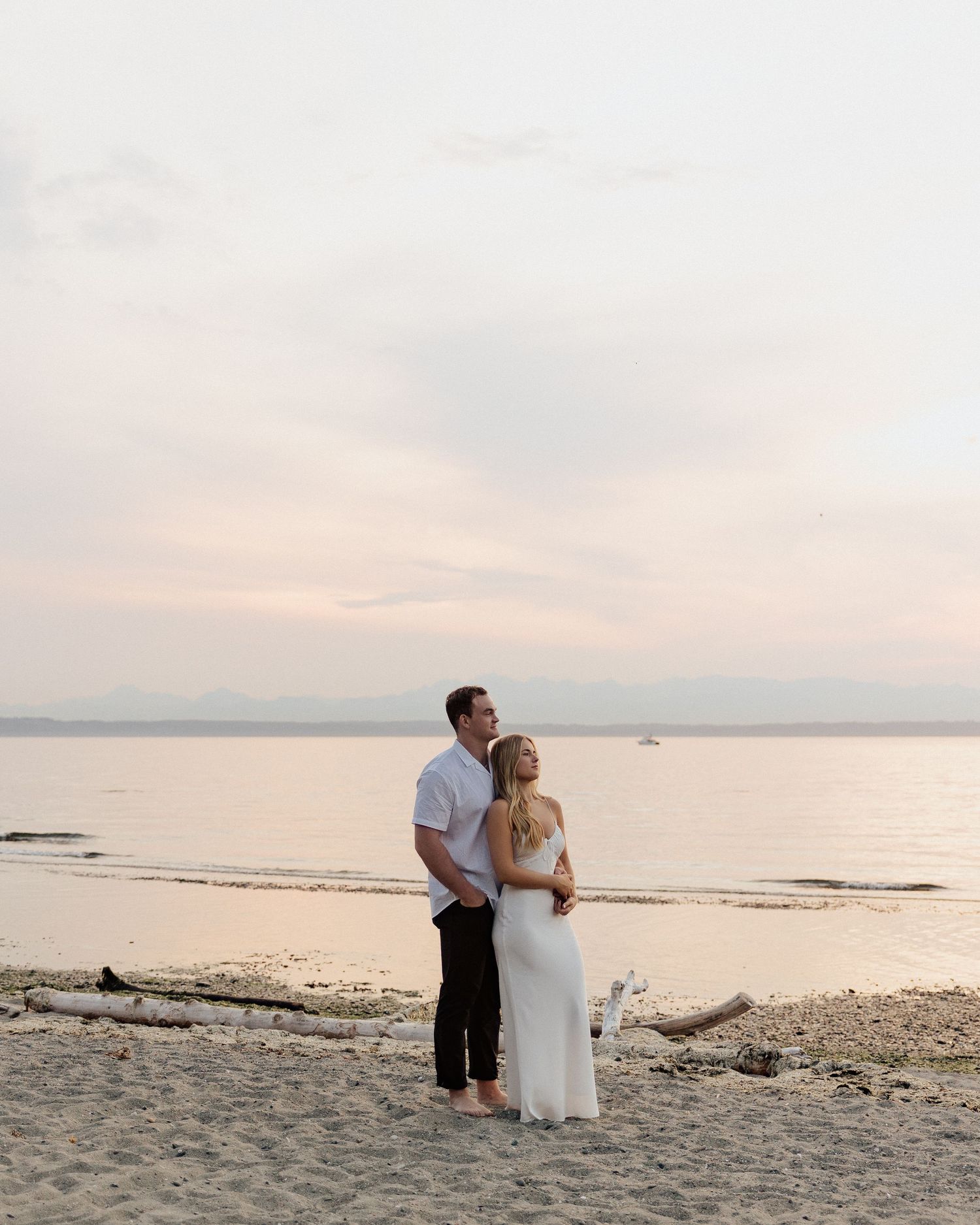 A romantic couple stands together on a beach at sunset with mountains in the distance.