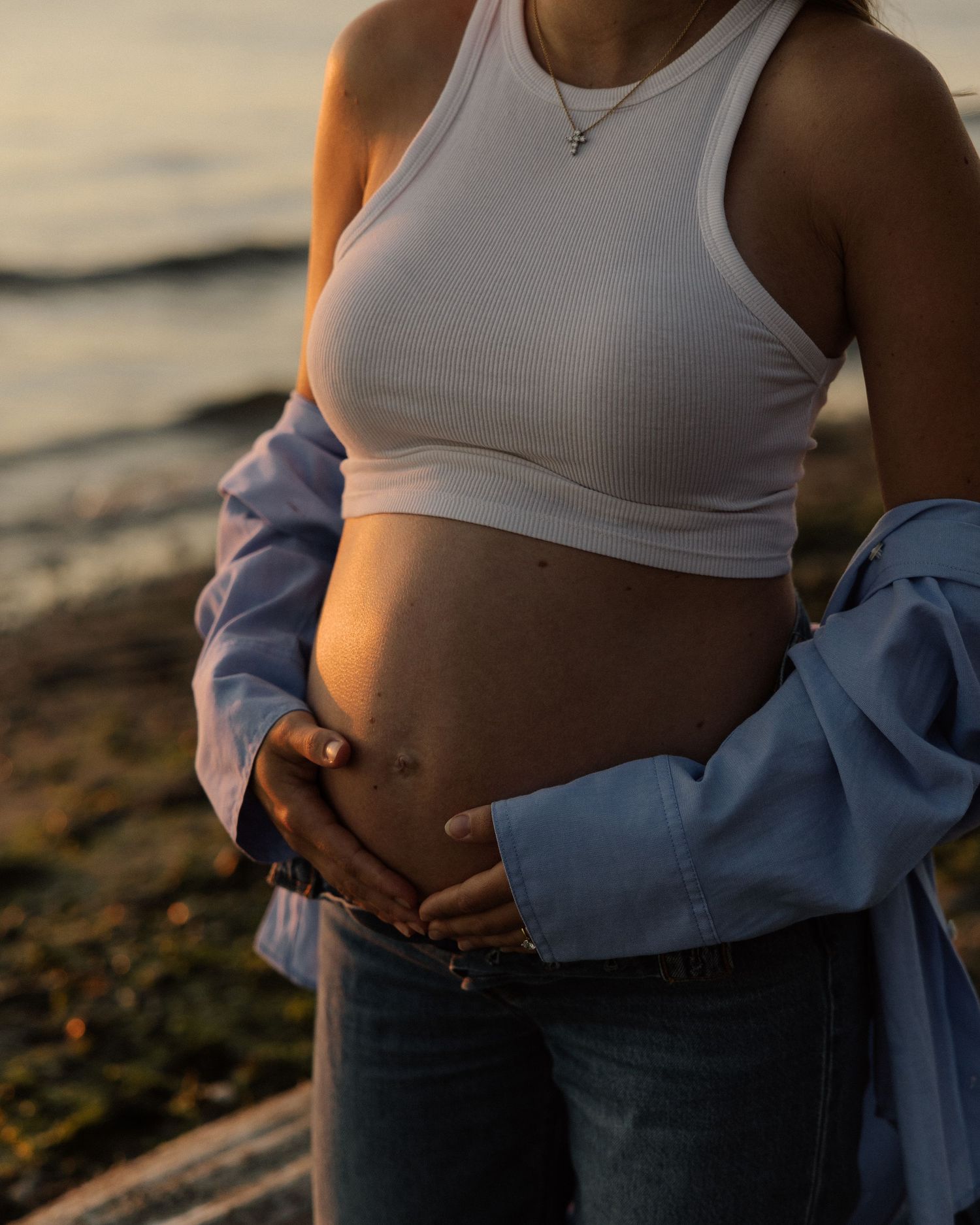 Close-up of pregnant belly with hands tenderly placed on it against sunset beach backdrop.