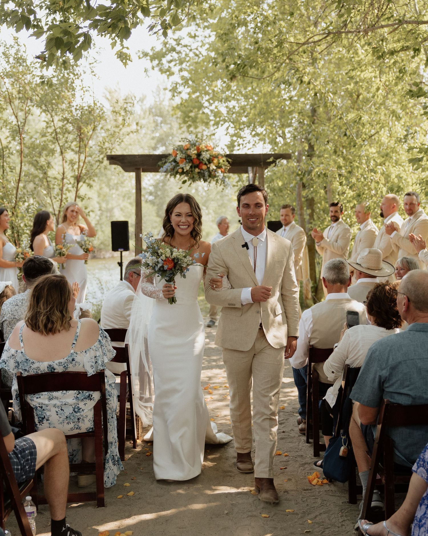 Couple walking down aisle after outdoor wedding ceremony while guests watch.