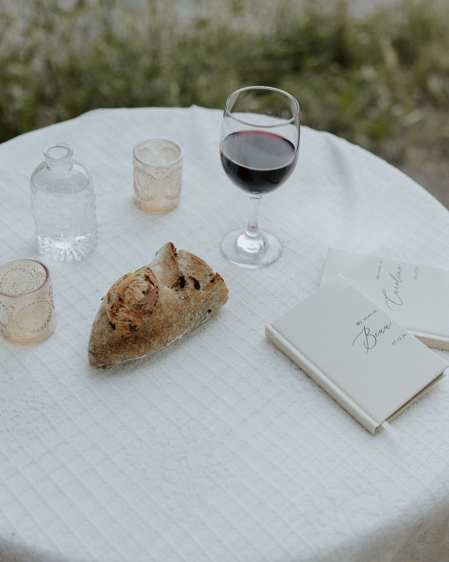 Glass of red wine, rustic bread, and a notebook on a white tablecloth in an outdoor setting.