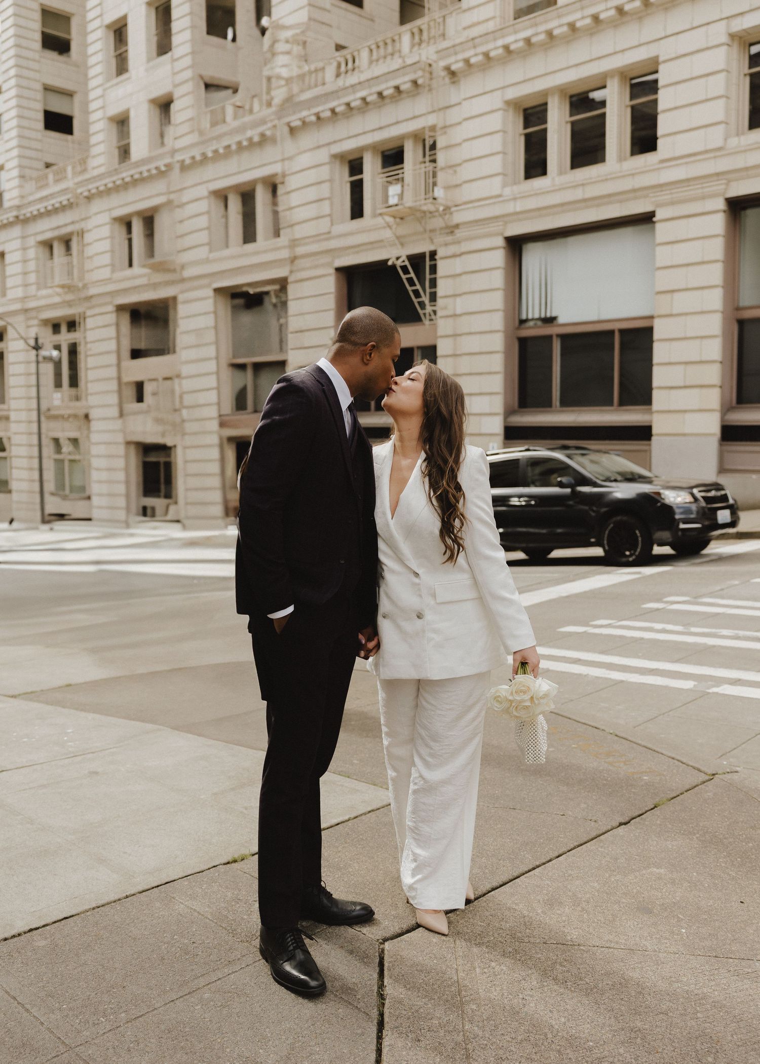 Newlyweds sharing a romantic kiss on a city sidewalk, groom in black suit and bride in elegant white pantsuit.