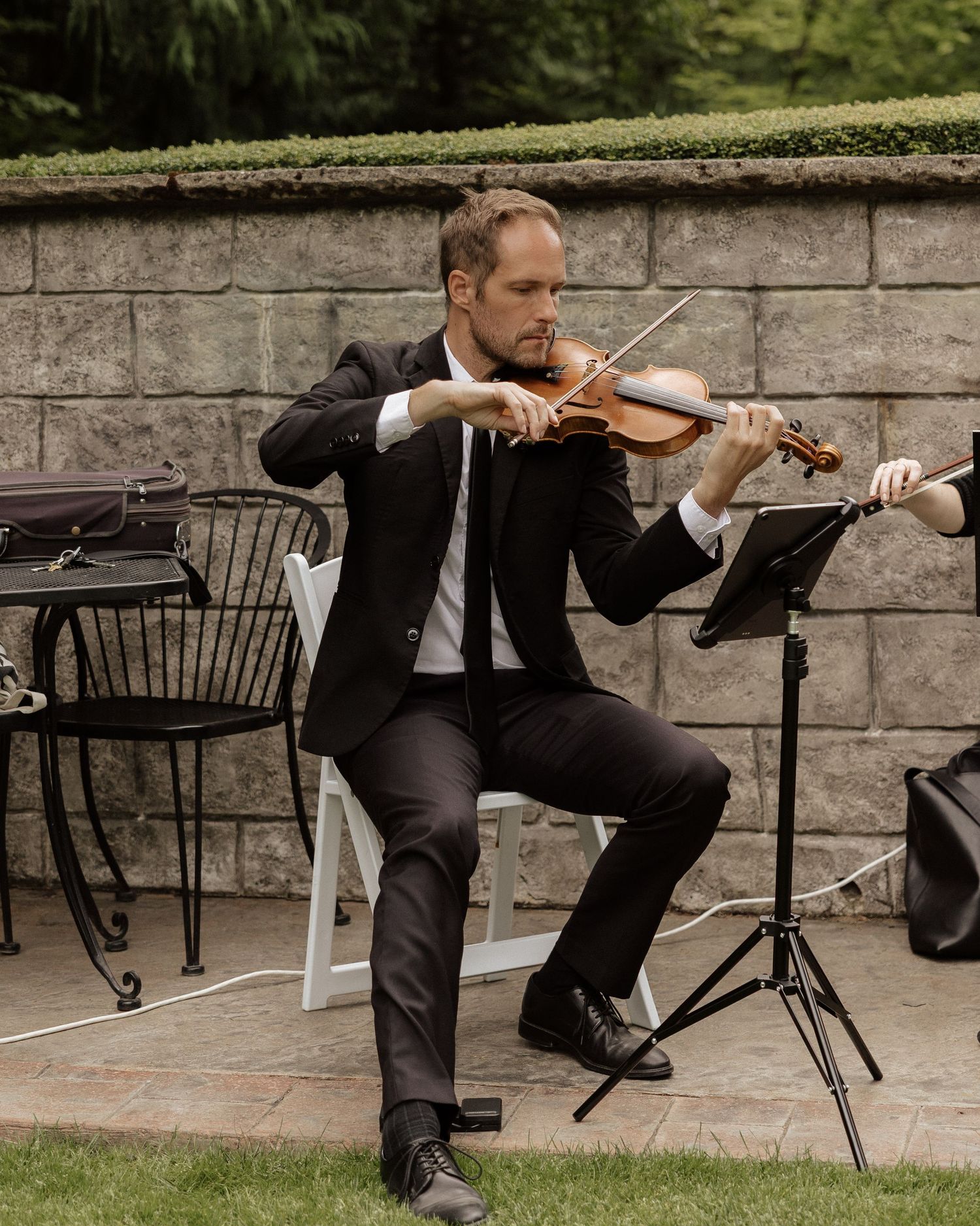 Male violinist in black suit performs outdoor wedding ceremony music on stone patio with music stand.