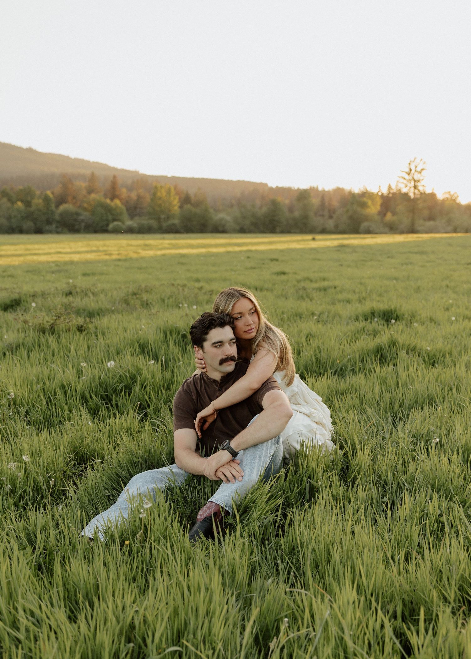 Couple sitting together in a lush green field at golden hour, with trees and hills in the background.