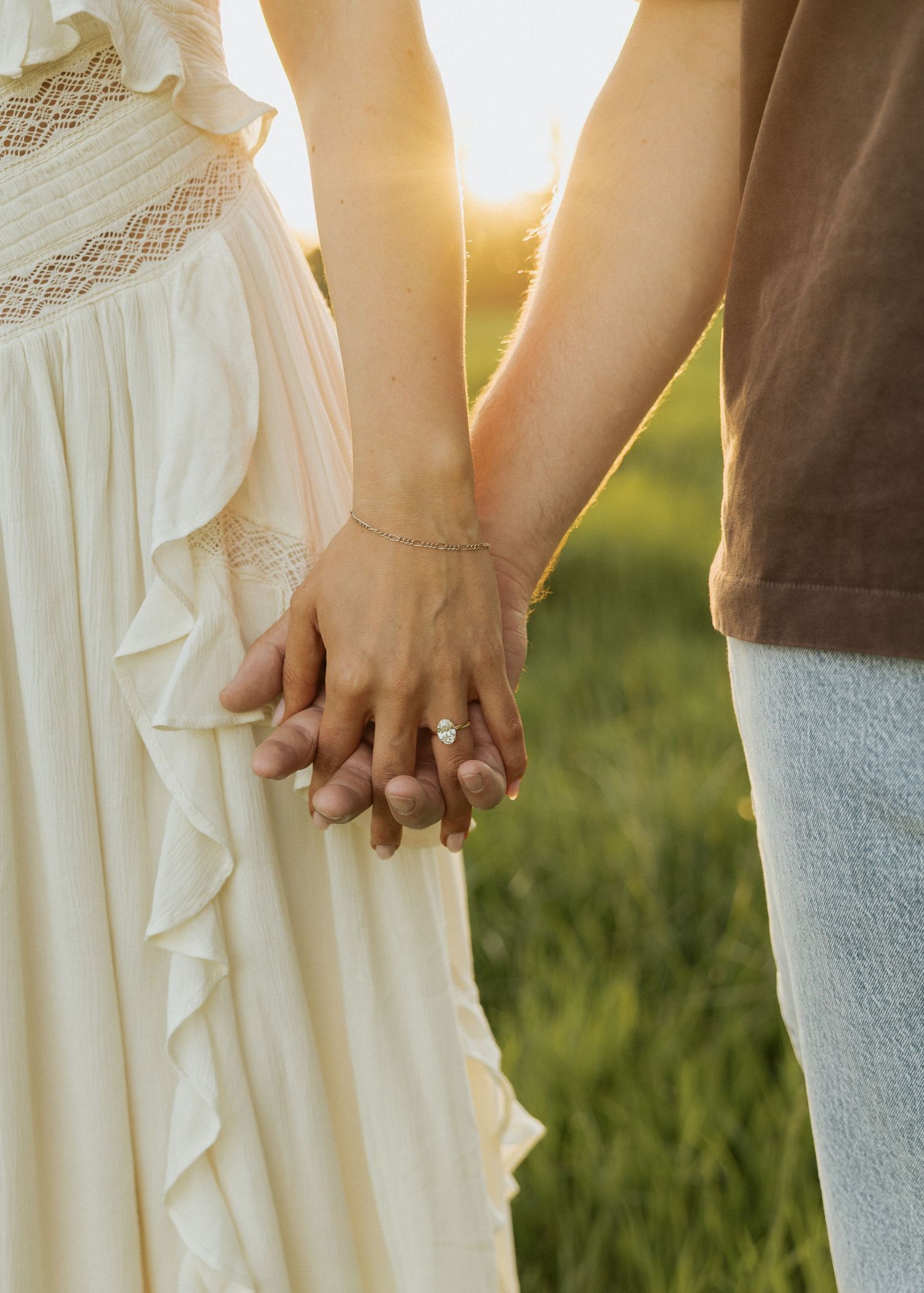 Couple holding hands at sunset, woman wearing engagement ring and white ruffled dress, golden hour outdoor photo.
