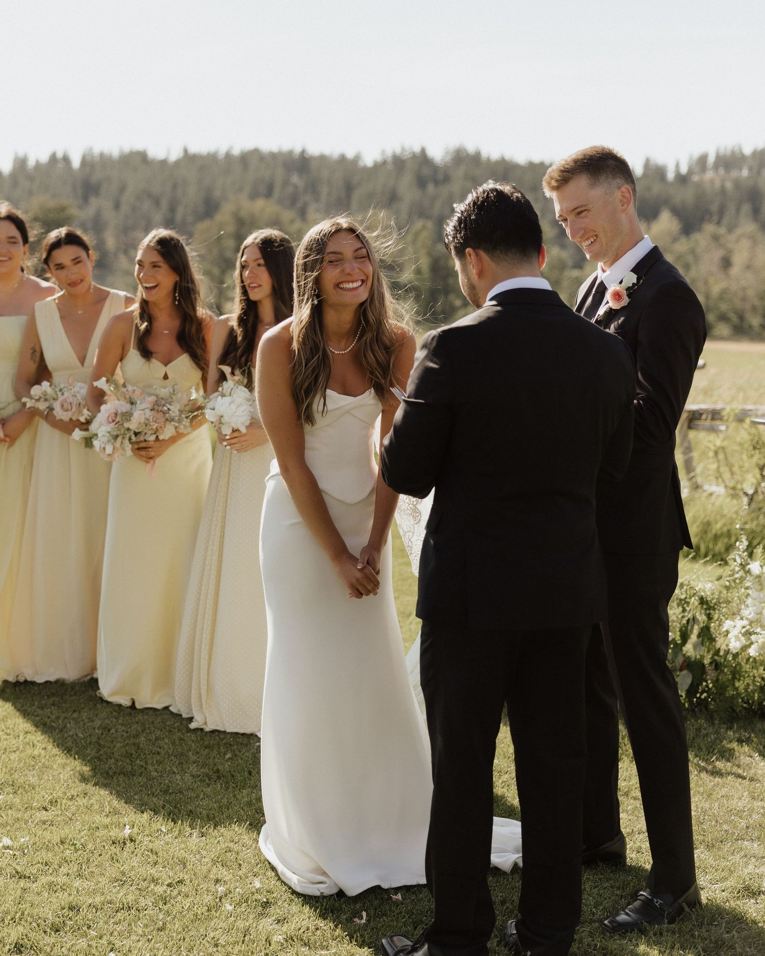 Bride smiling during outdoor wedding ceremony with bridesmaids in yellow dresses and groom in black suit.