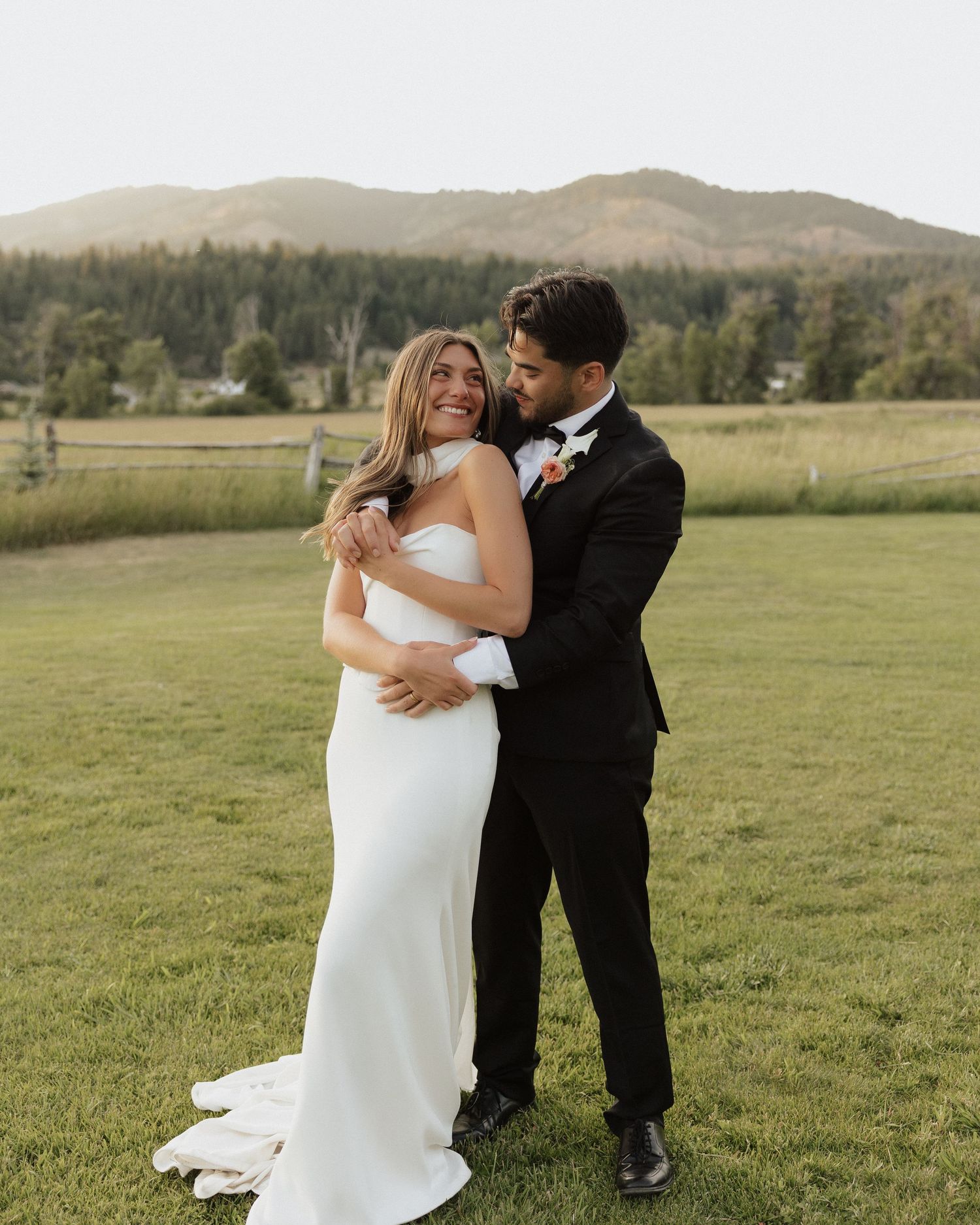 A bride in a white gown and groom in a black tuxedo embrace joyfully on a green lawn with mountain scenery.