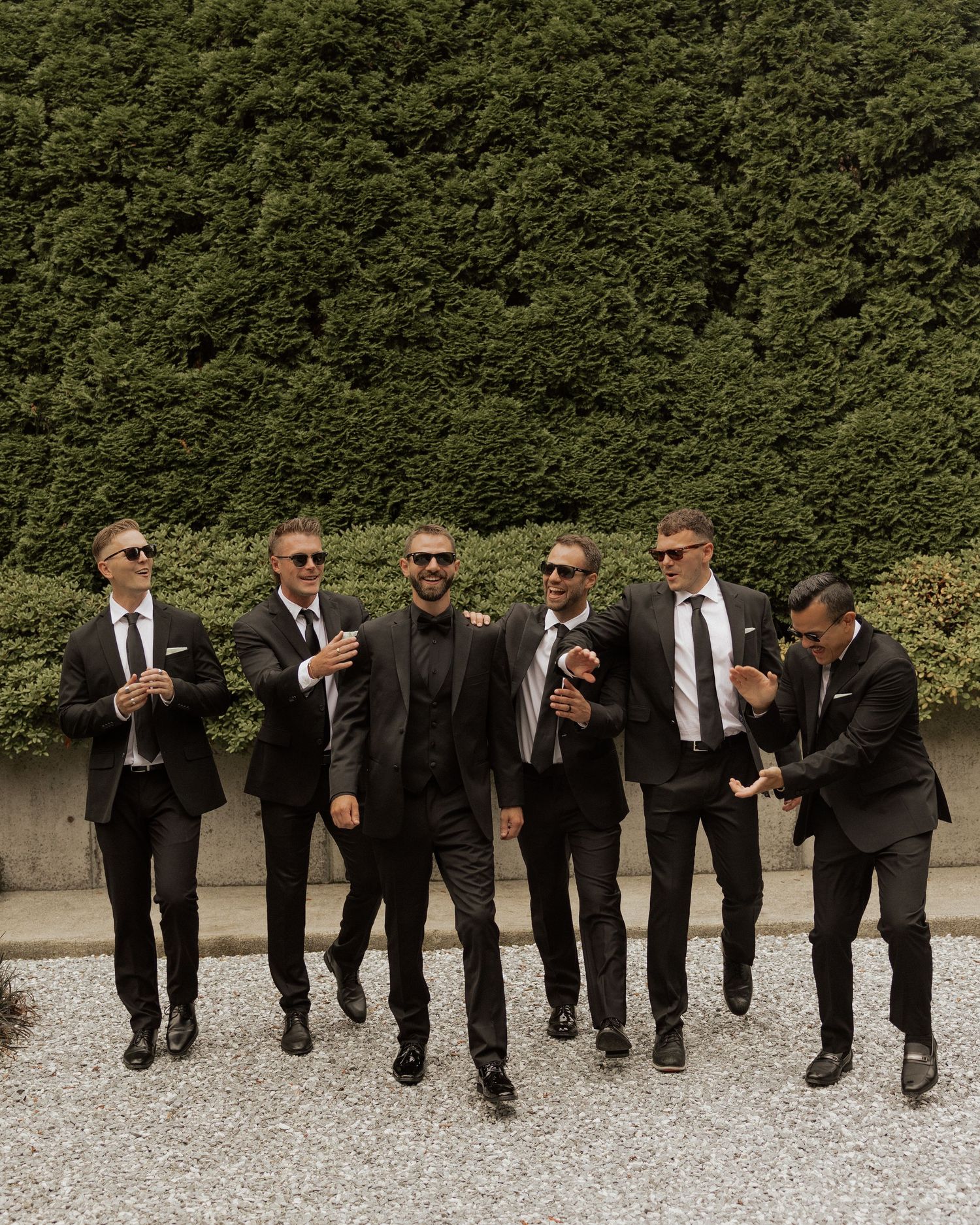 Six groomsmen in black suits and sunglasses laugh and walk together outdoors at a wedding.