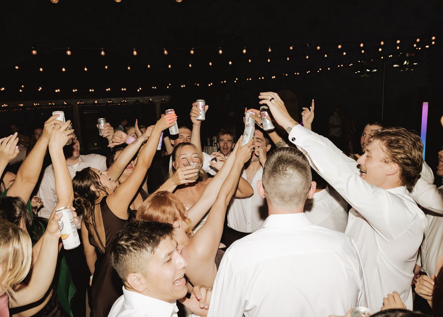 Crowded wedding reception dance floor with guests celebrating under warm string lights at night.