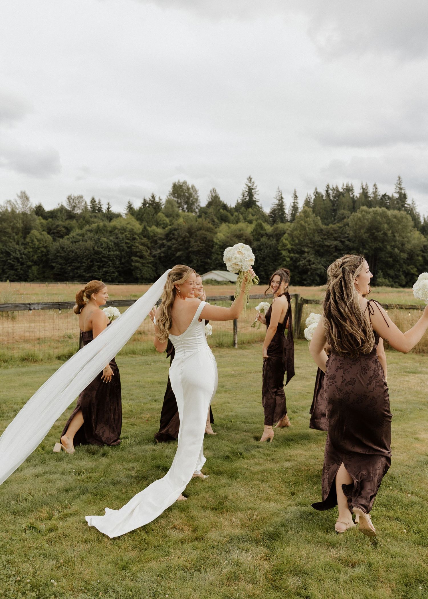 Bride in white gown and veil walks with bridesmaids in brown dresses holding bouquets on green lawn.
