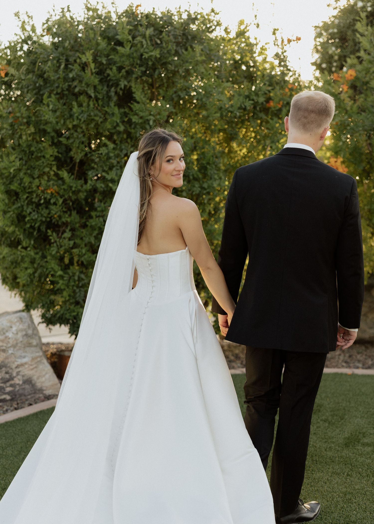 Bride in strapless white gown and veil holds hands with groom in black suit, walking through lush garden.