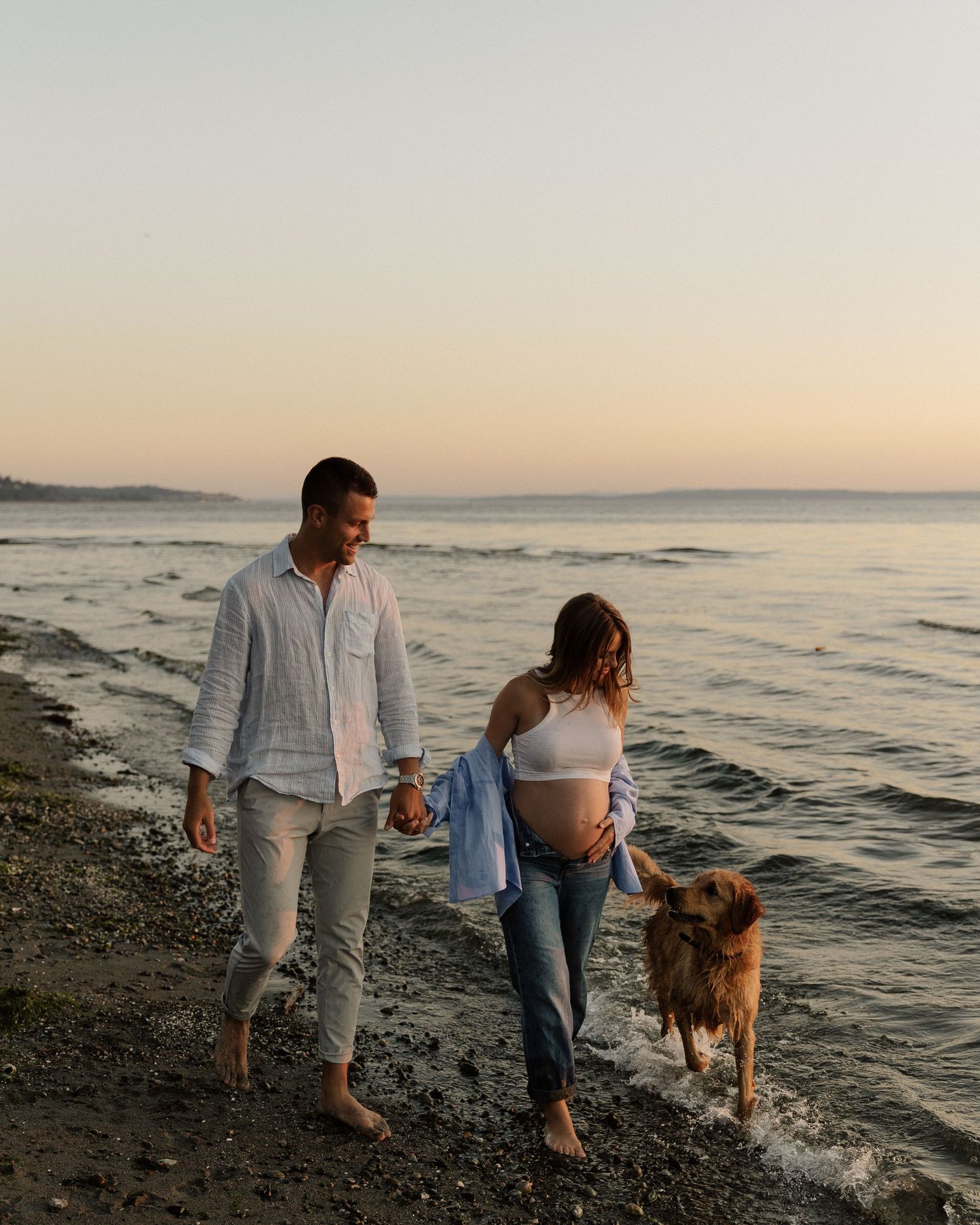 A couple walks their dog along a scenic beach shoreline at sunset while dressed in casual summer attire.