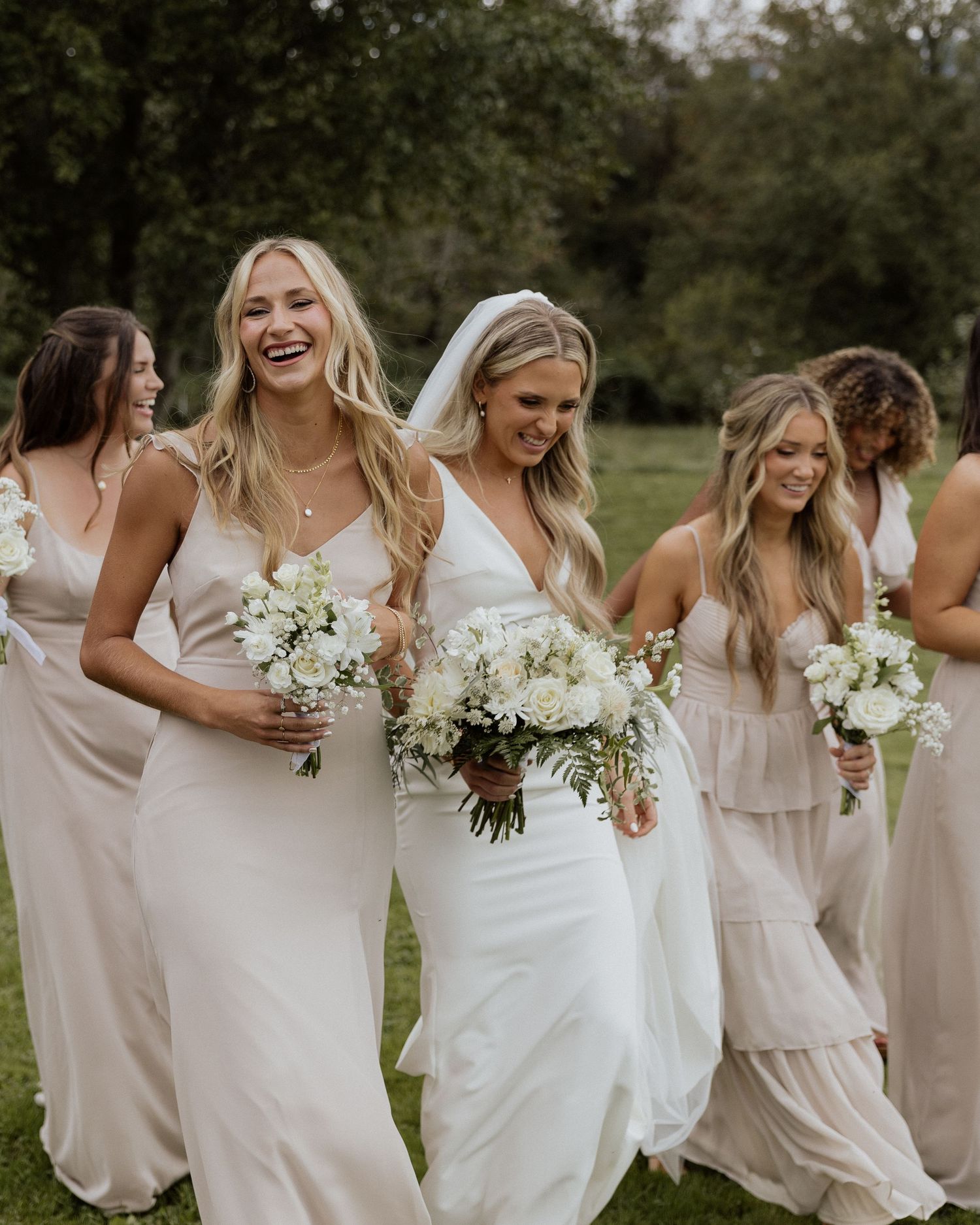 Bridesmaids in blush dresses carrying white bouquets share joyful laughter together outdoors.