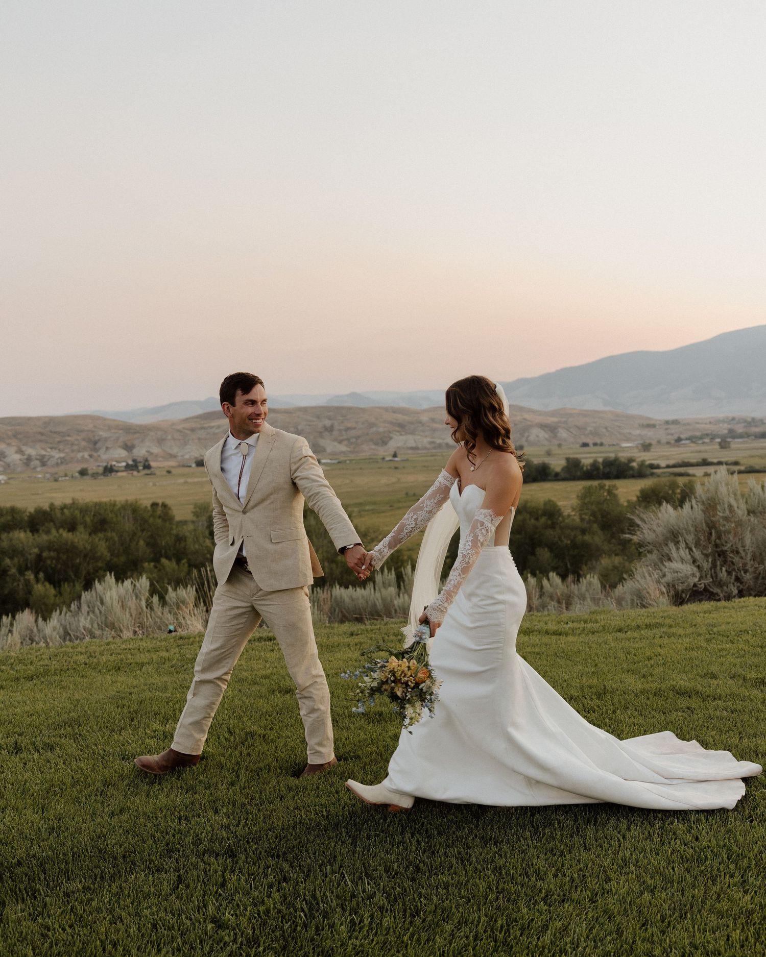 Newlyweds walk hand in hand across hilltop meadow at sunset, bride in white gown and groom in beige suit.