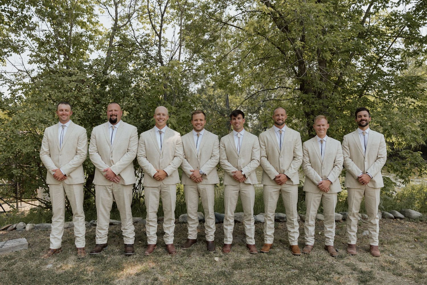 Wedding party of groomsmen in matching beige suits standing in a line outdoors.