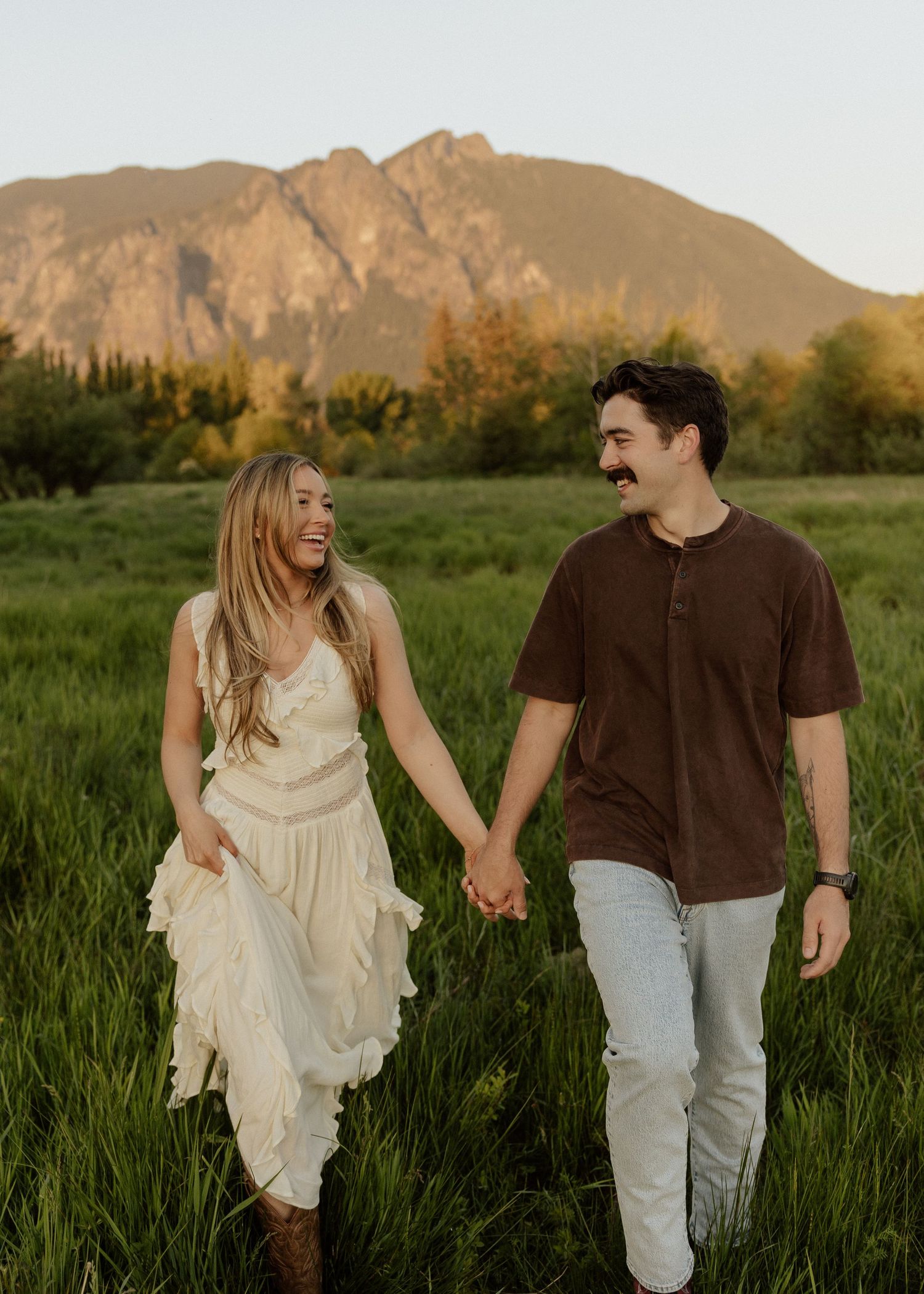 A couple holds hands and smiles while walking through a green field with a mountain backdrop at golden hour.