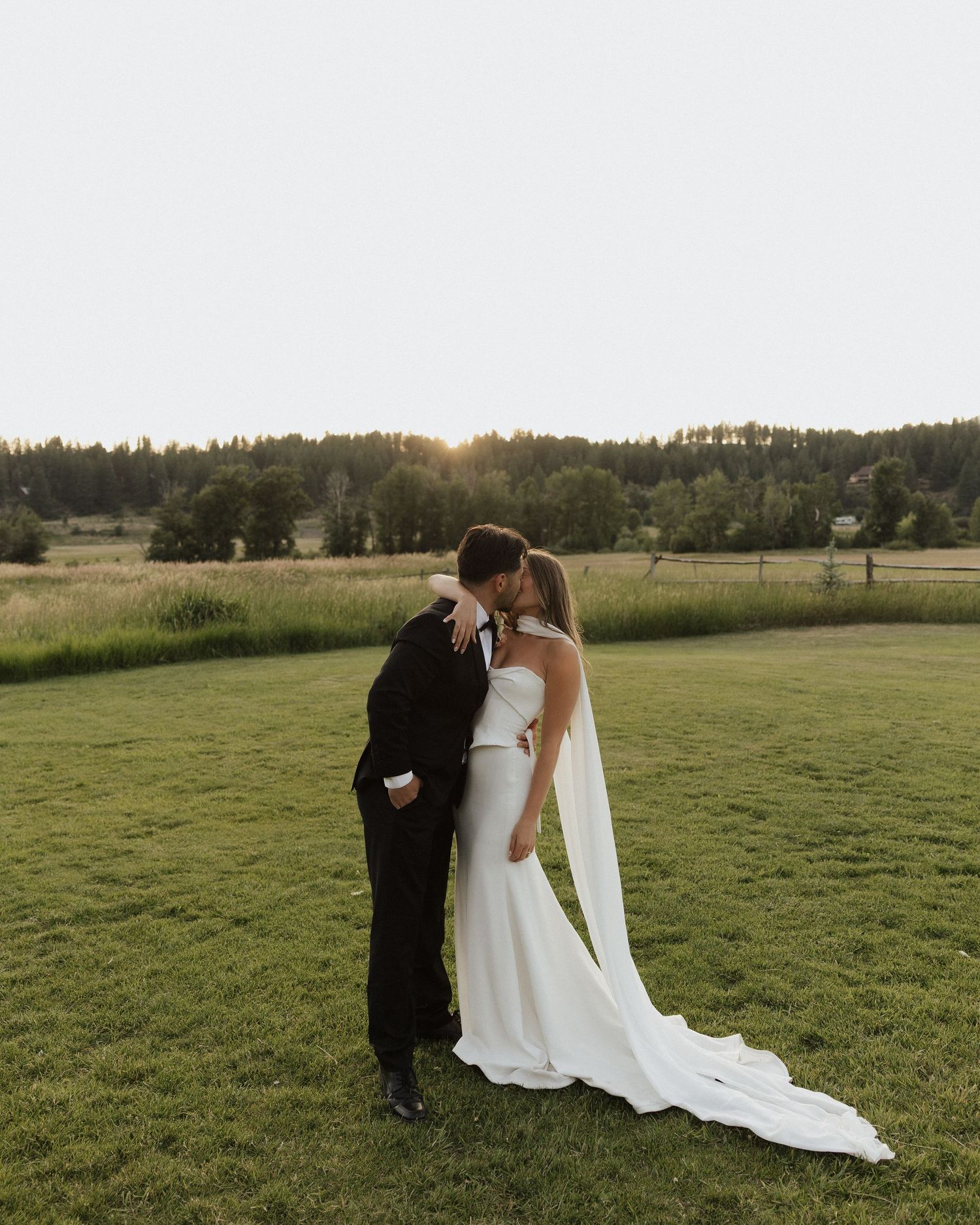 Bride and groom share romantic sunset kiss on green lawn at outdoor countryside wedding.