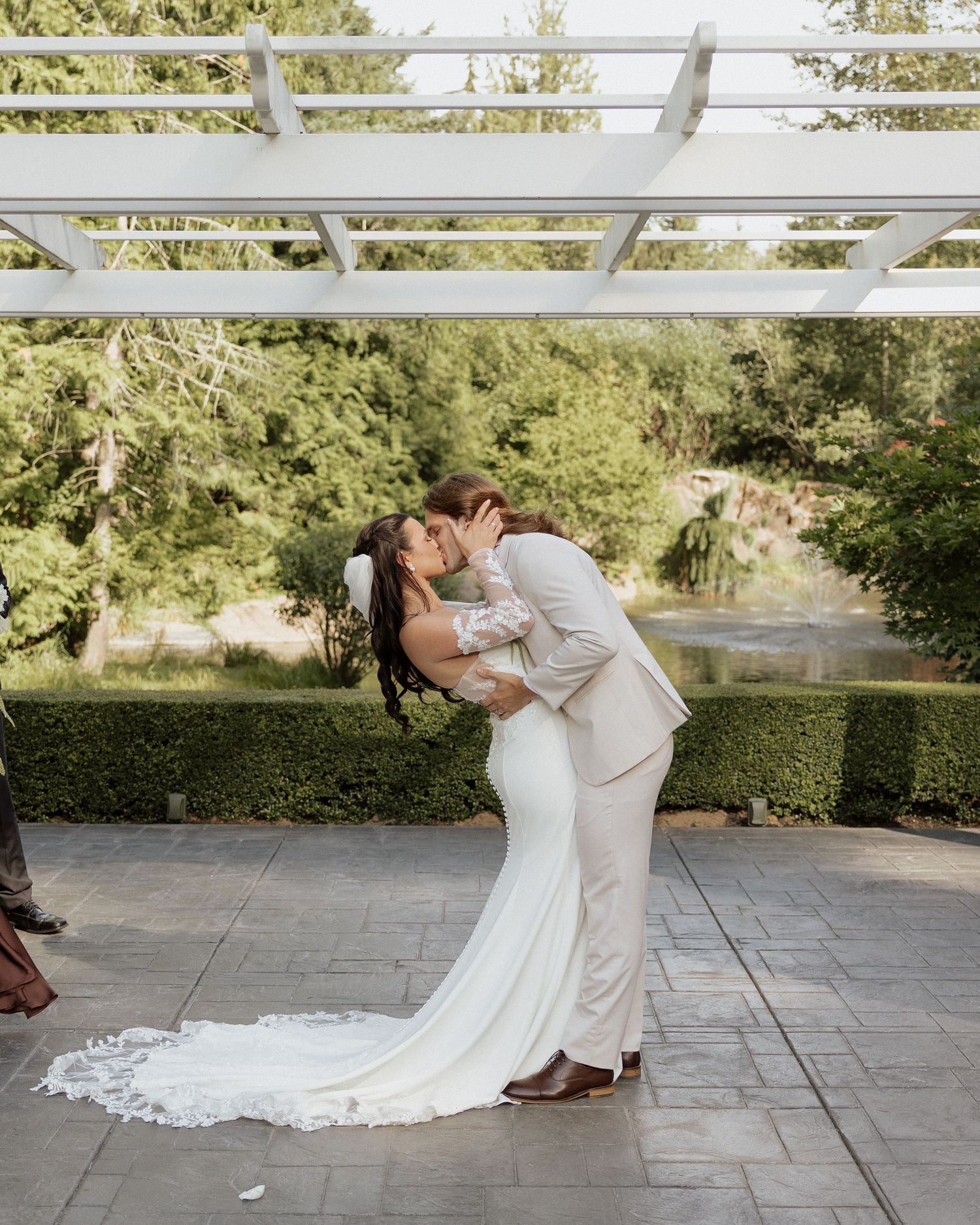 Bride and groom share a romantic first kiss under a white pergola with lush green garden backdrop on their wedding day.