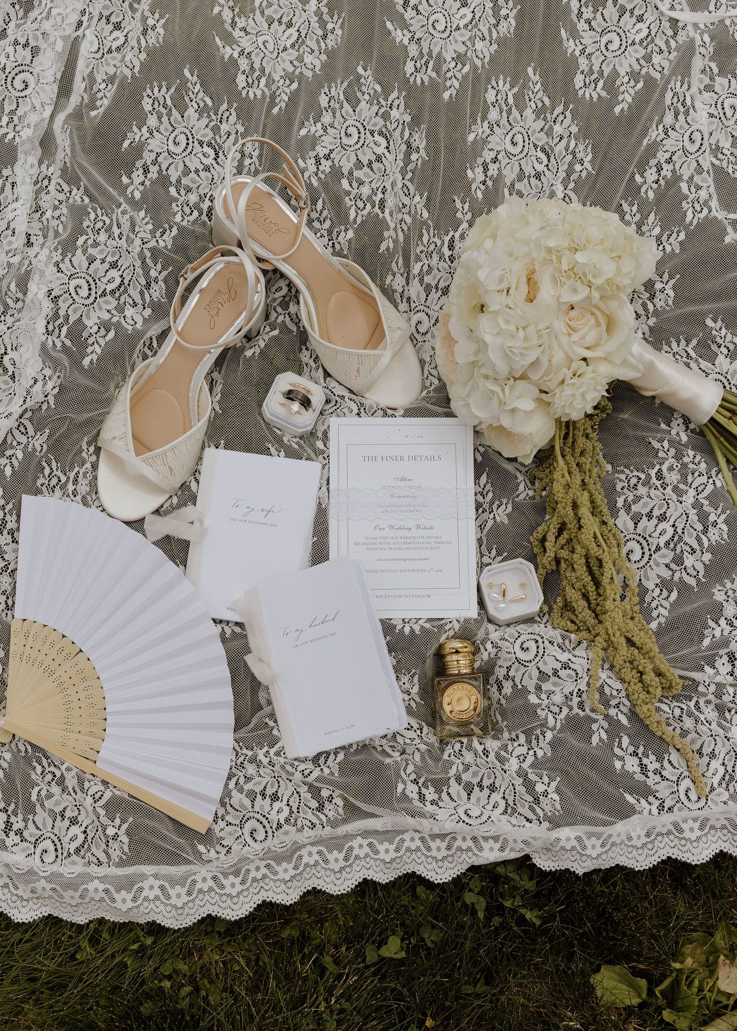 Flat lay of wedding details including ivory heels, white hydrangea bouquet, stationery, fan, and gold perfume bottle.