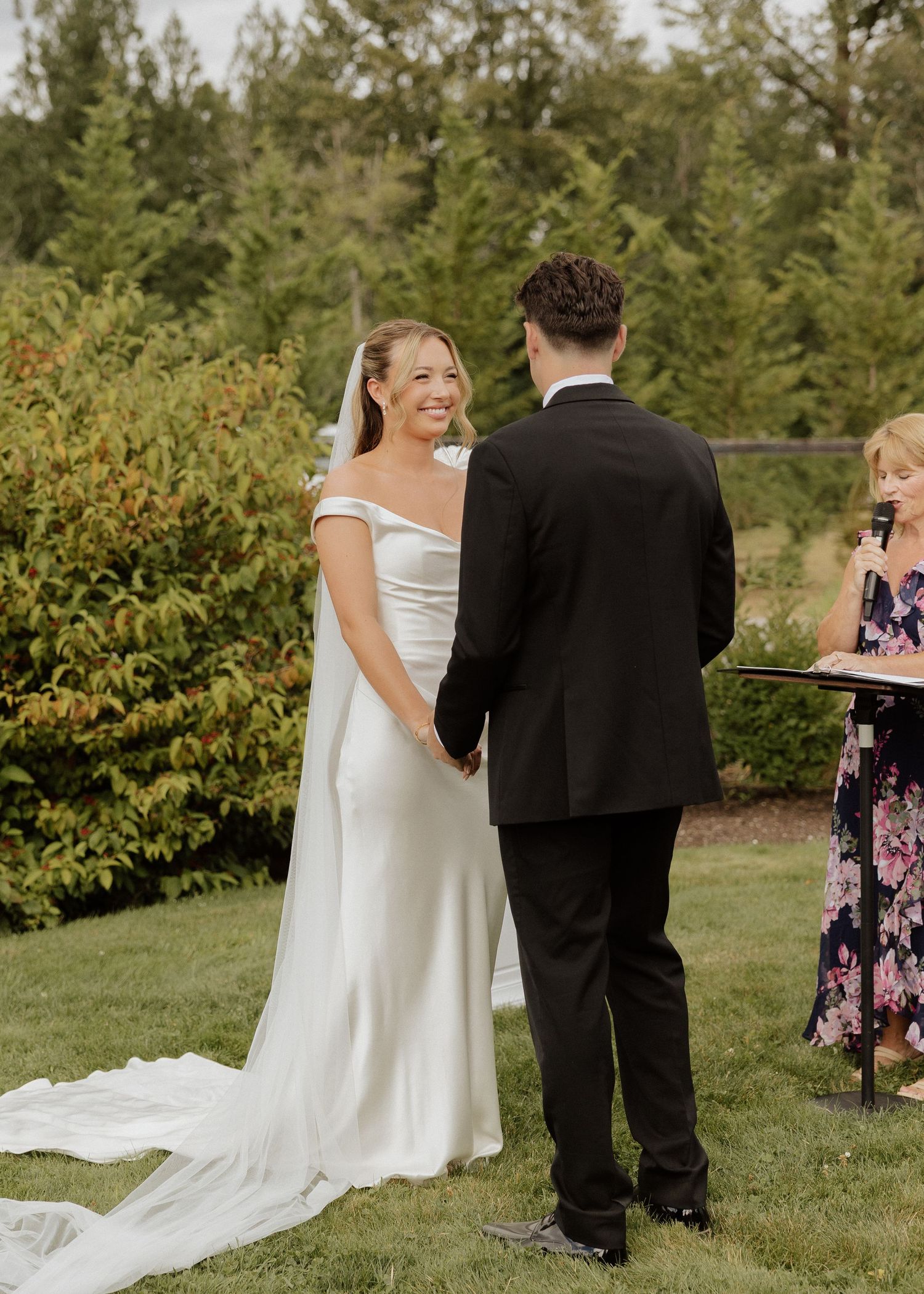 Smiling bride in off-shoulder white gown holds hands with groom in black suit during outdoor wedding ceremony.