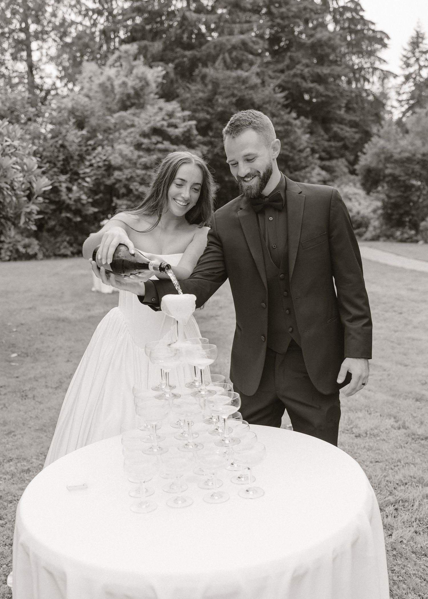 Bride and groom pouring champagne tower at outdoor wedding reception in black and white photo.