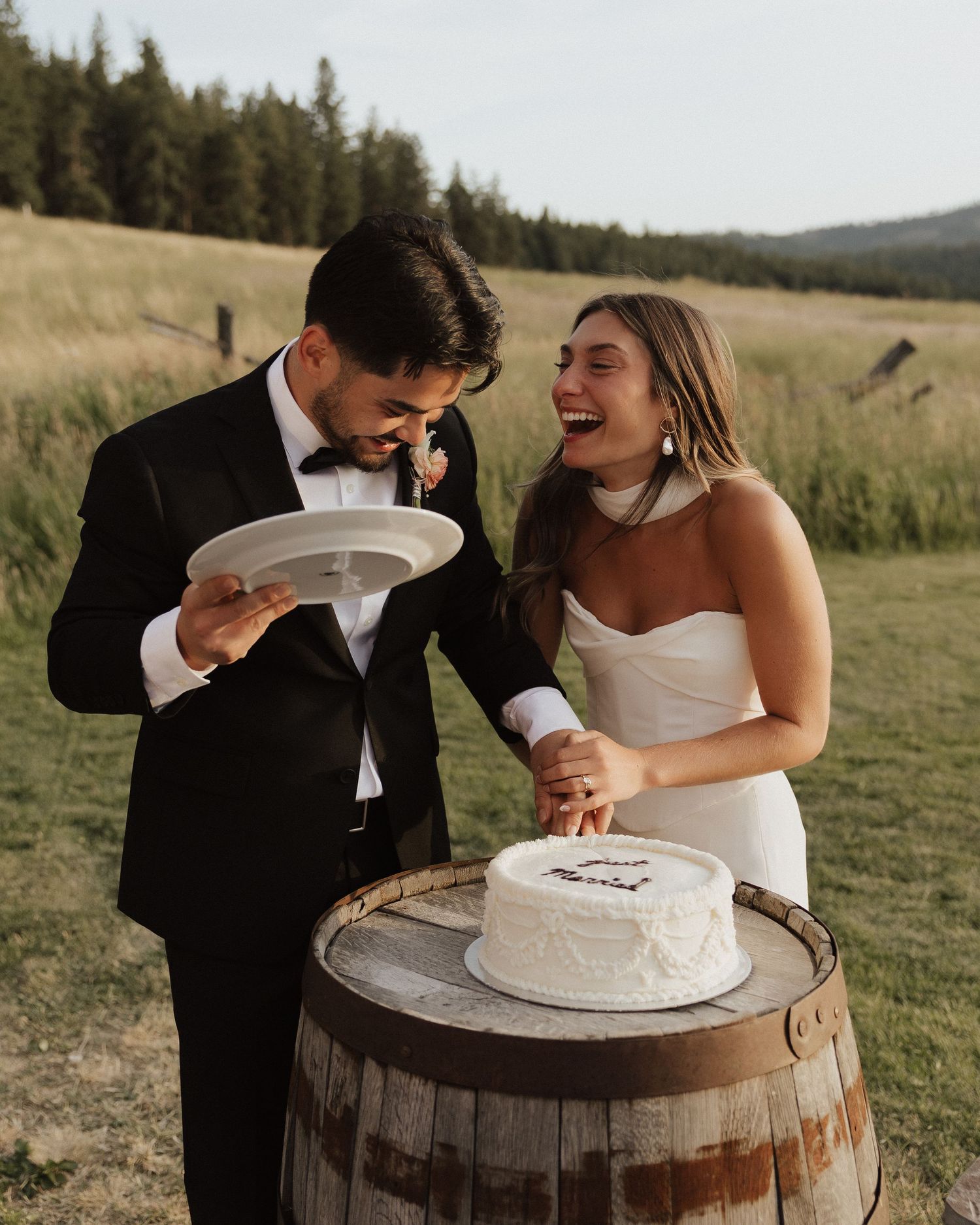 Newlyweds cutting their white wedding cake displayed on a rustic wooden barrel outdoors.