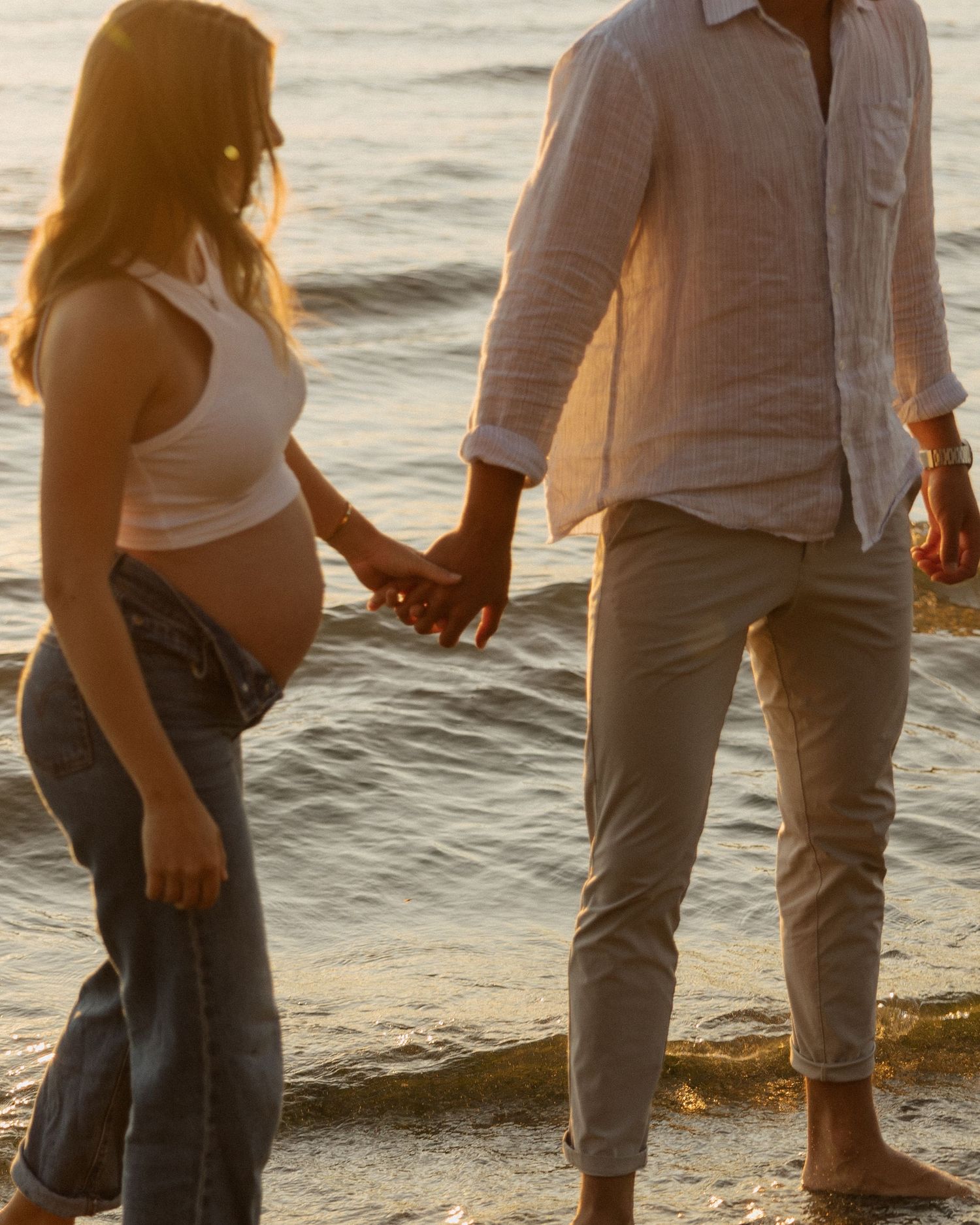Expecting couple holds hands while walking along shoreline at golden hour.