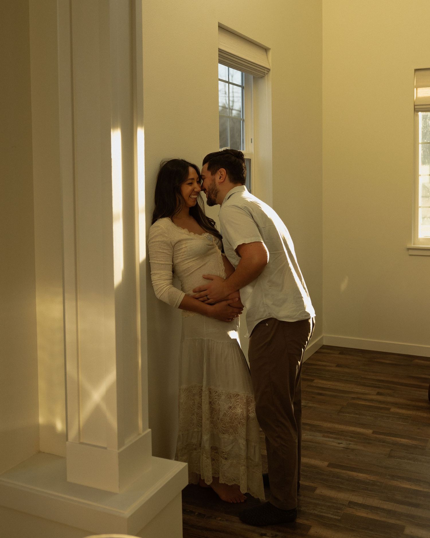 Sunlit maternity photo with couple embracing near a window.