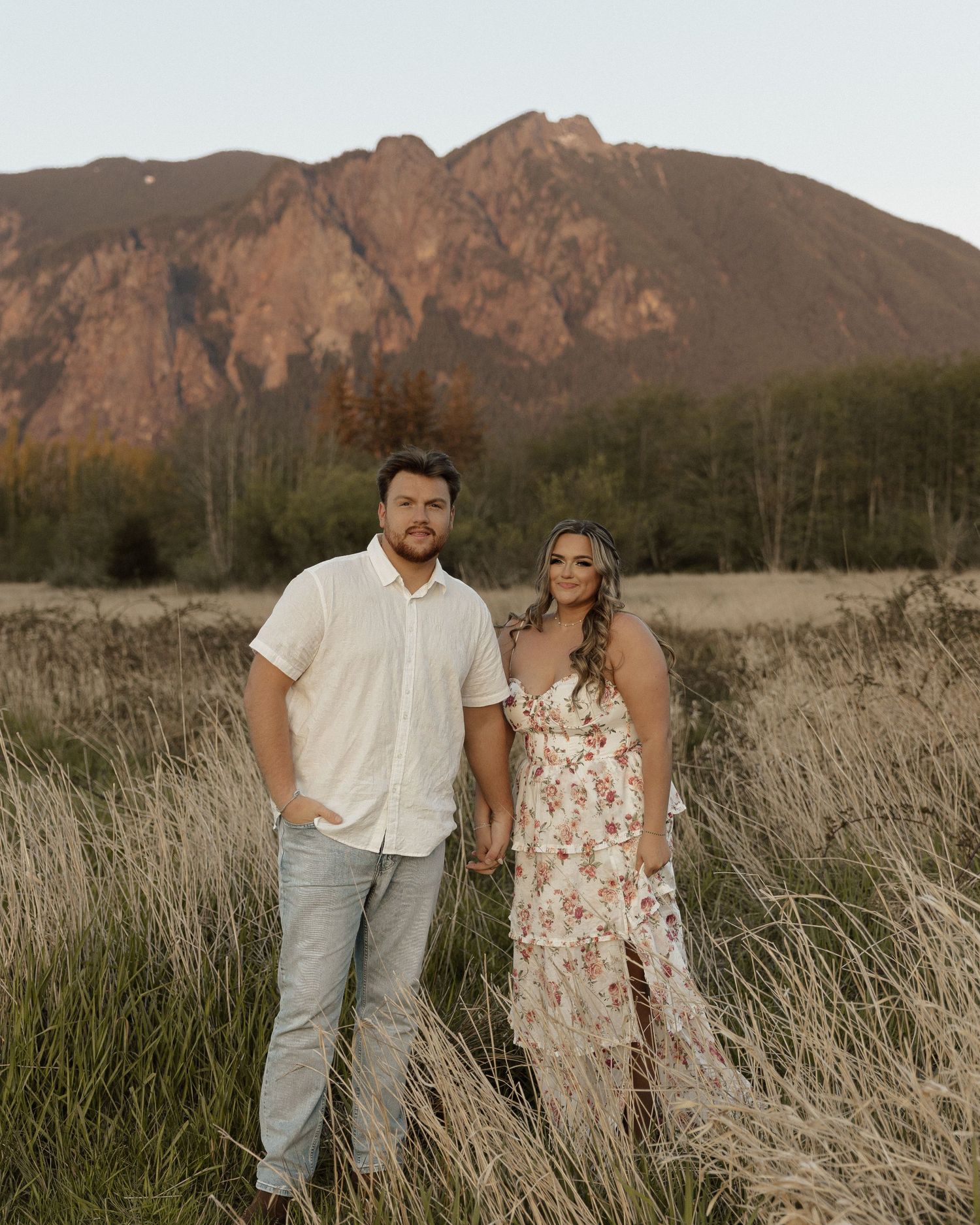 Couple standing in tall grass field with majestic mountain backdrop during sunset.