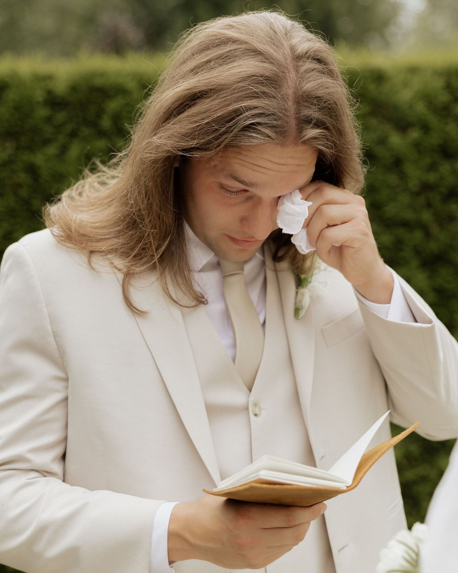 Emotional groom in ivory suit wiping tears while reading vows at outdoor wedding ceremony.
