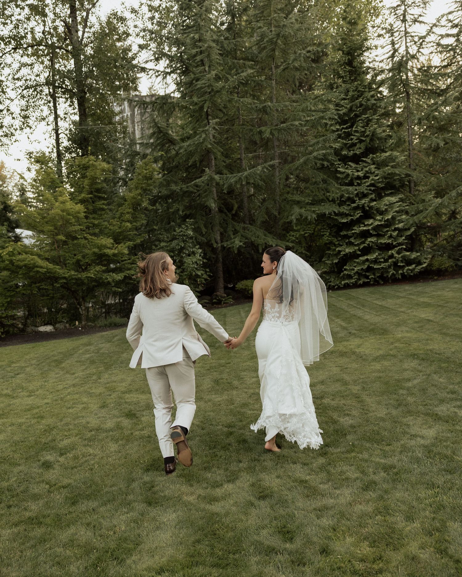 Bride in white lace gown and groom in beige suit holding hands while walking across green lawn near tall trees.