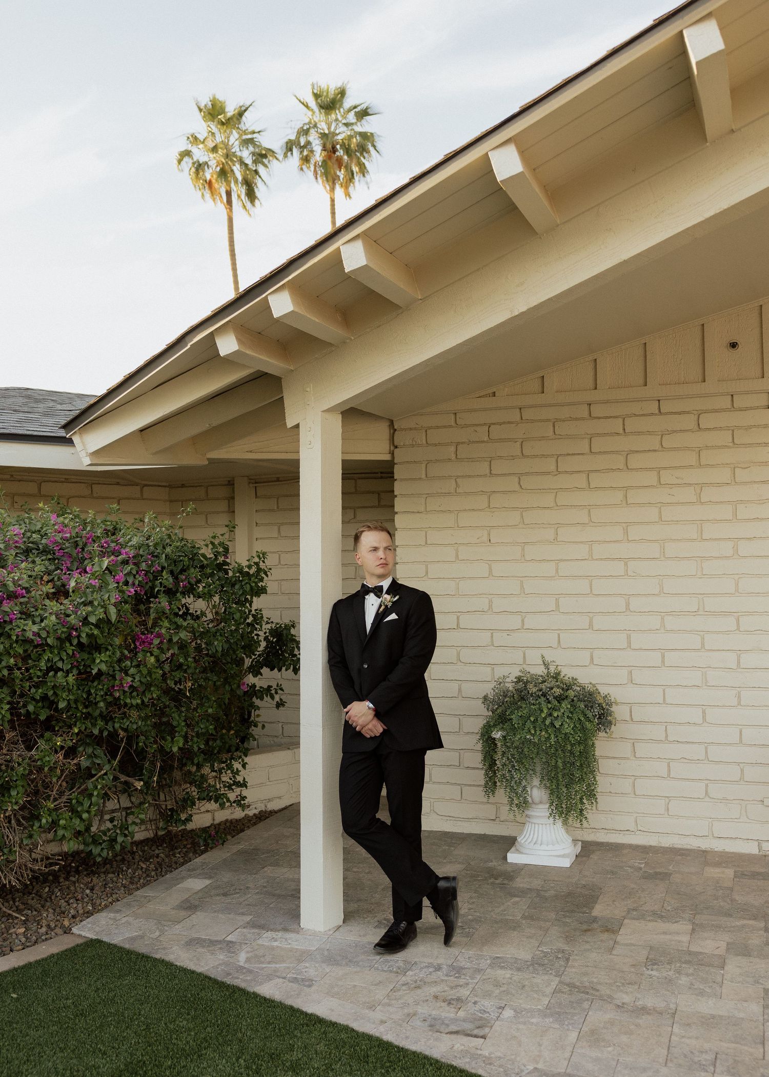 Groom in black tuxedo leaning against white brick pillar on patio with palm trees and lush greenery in background.