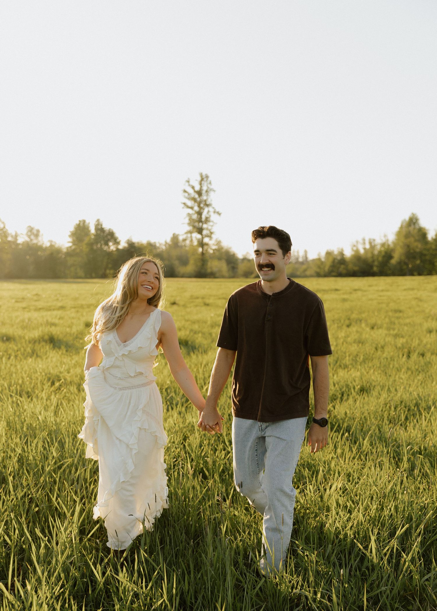 A couple holds hands and walks through a lush green field at golden hour, woman in white dress, man in dark shirt.