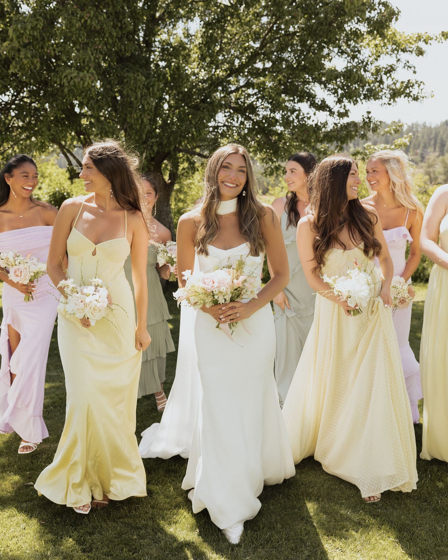 Smiling bride in white dress walks with bridesmaids in yellow and neutral gowns holding bouquets outside.