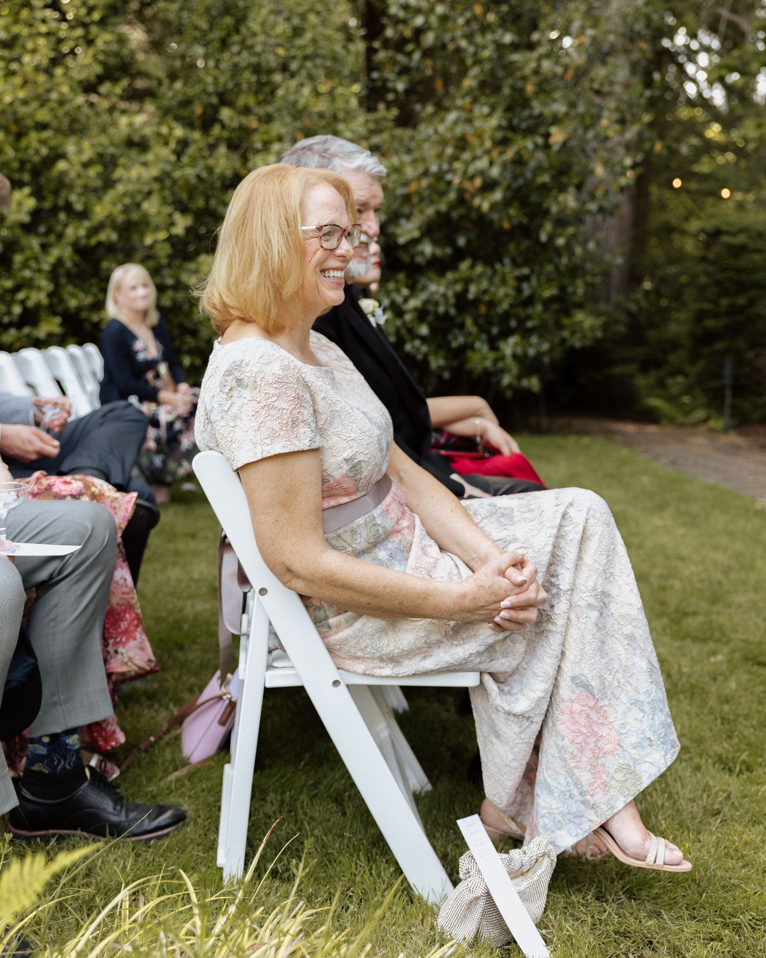 Smiling woman in floral dress seated in white folding chair at outdoor wedding ceremony on green lawn.