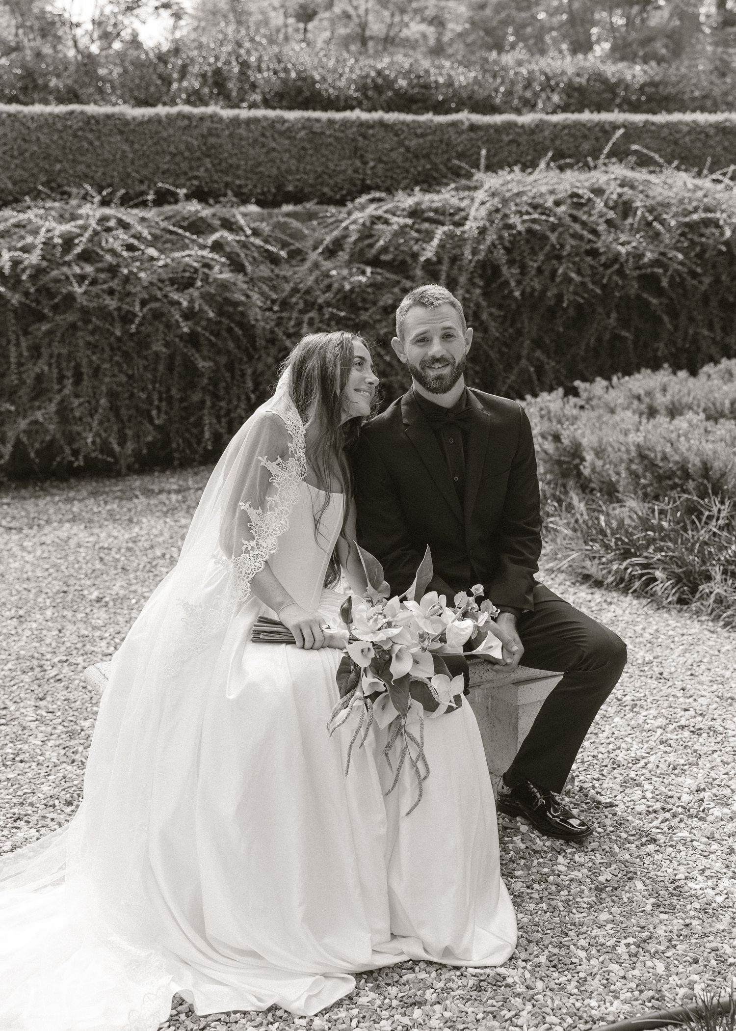 Black and white photo of bride and groom sitting together in garden, bride holding bouquet in wedding dress with veil.