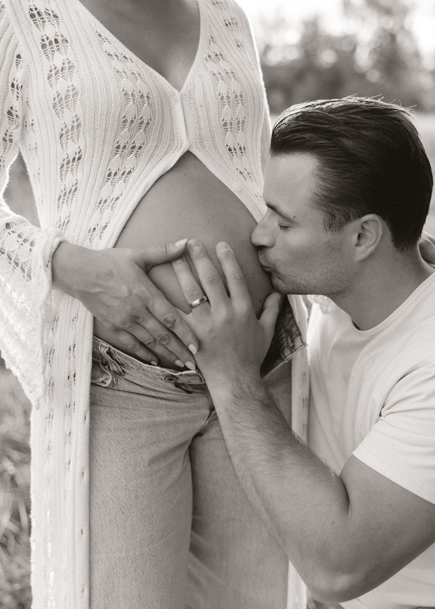 Black and white photo of man kissing pregnant woman's baby bump outdoors during maternity photoshoot.