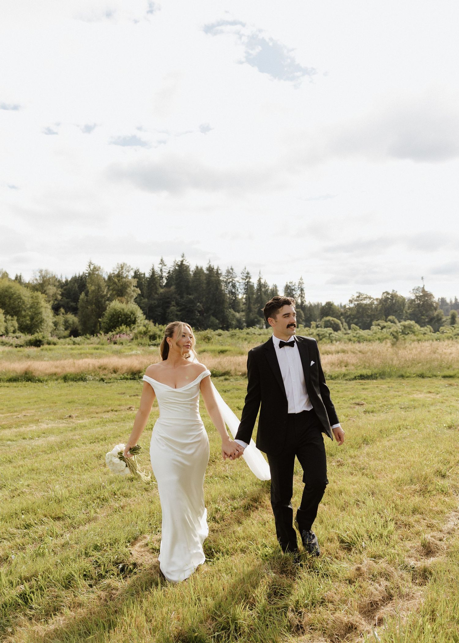 Bride and groom walk hand in hand through a lush green meadow on their wedding day.