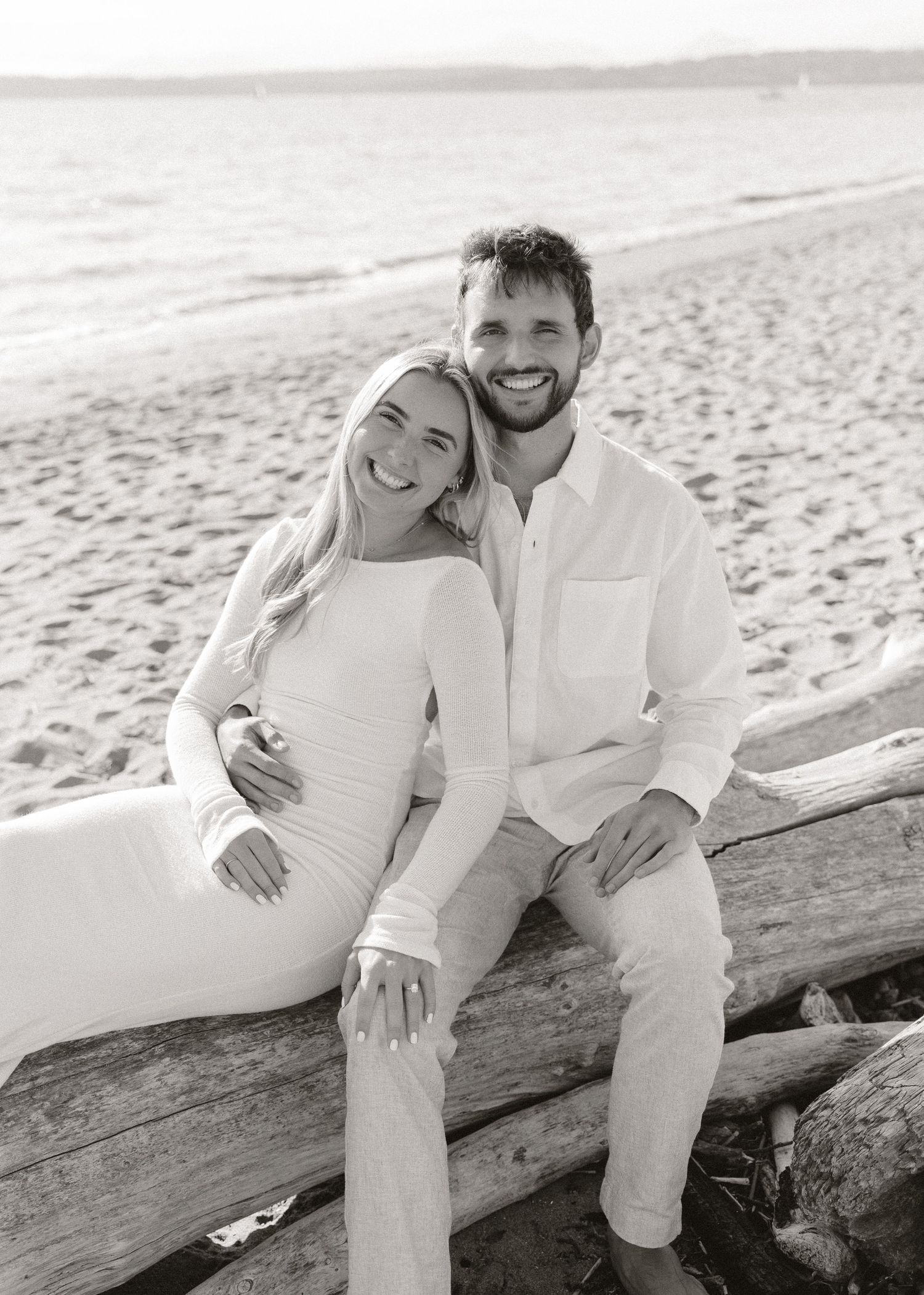 Black and white beach photo of smiling couple sitting on driftwood log, woman in white dress, man in white shirt.