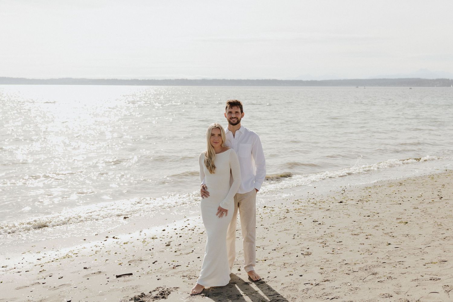 Couple in white outfits posing on a sandy beach with calm ocean water in the background on an overcast day.