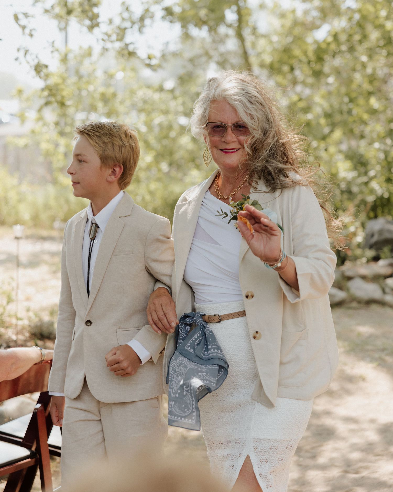 A young boy and older woman in matching light colored outfits walk arm in arm at outdoor wedding.