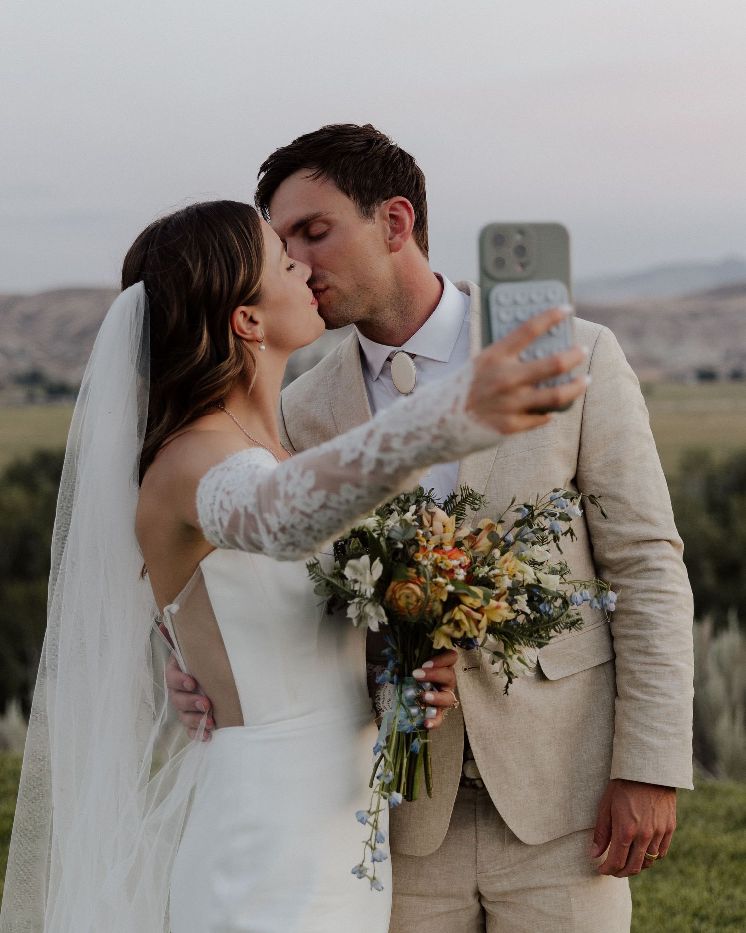 Newlyweds sharing a kiss while taking selfie, bride holding colorful bouquet.