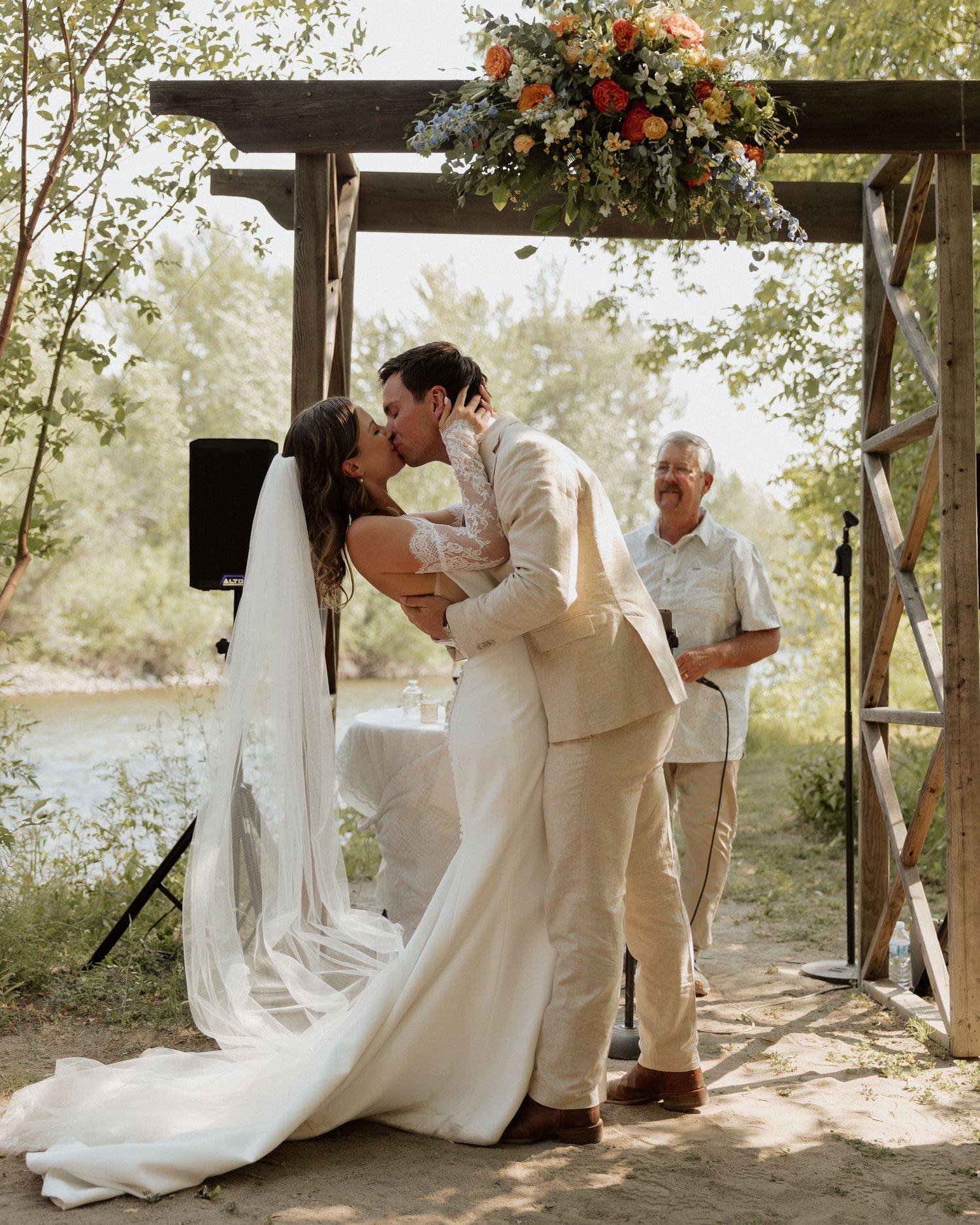 A romantic kiss under a wooden arch decorated with floral arrangements during an outdoor wedding ceremony.