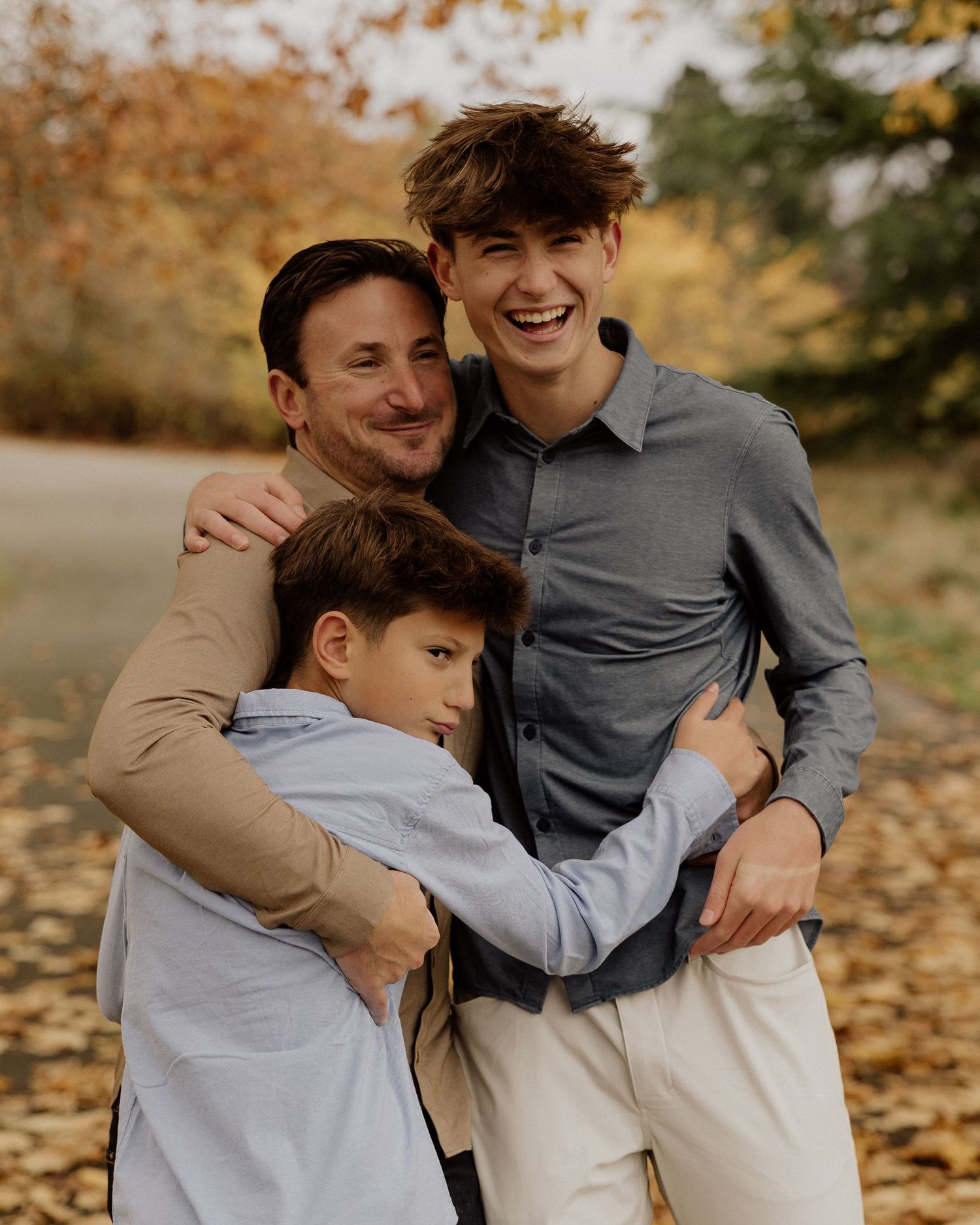 Three people embrace and laugh together on a fall day surrounded by golden foliage.